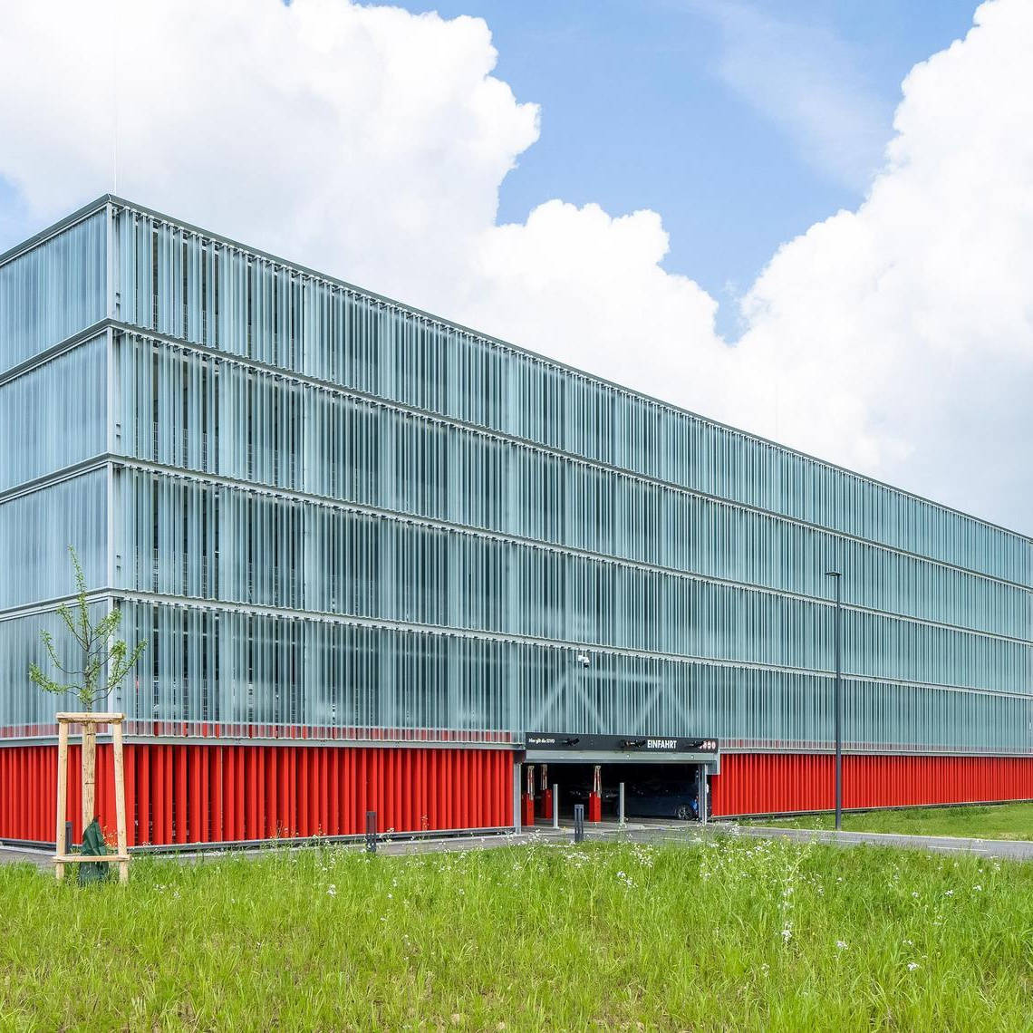 Modernes Parkhaus mit Glasfassade und roten Elementen, umgeben von Grünfläche und bewölktem Himmel.
