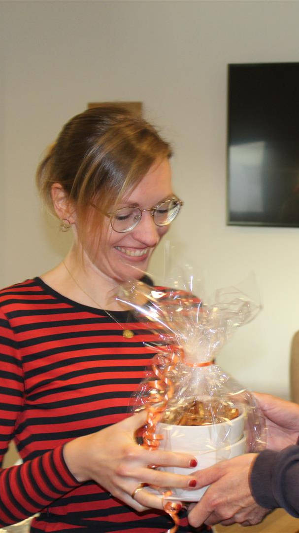 Frau in gestreiftem Oberteil erhält lächelnd ein Geschenk im Büro, während zwei Personen im Hintergrund zusehen.