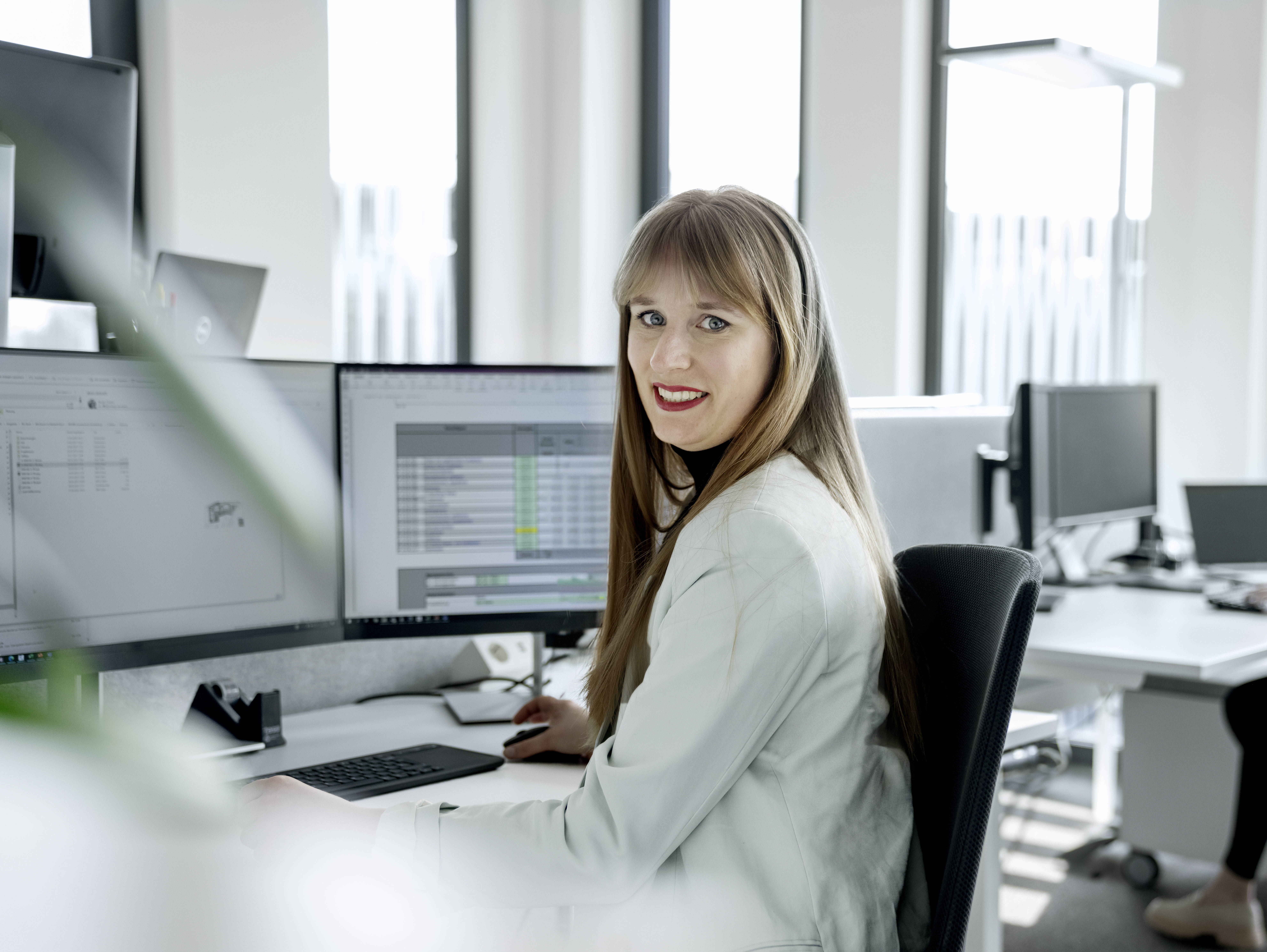 Frau im Büro an einem Schreibtisch mit mehreren Monitoren, lächelt in die Kamera. Im Hintergrund arbeitet eine Kollegin.