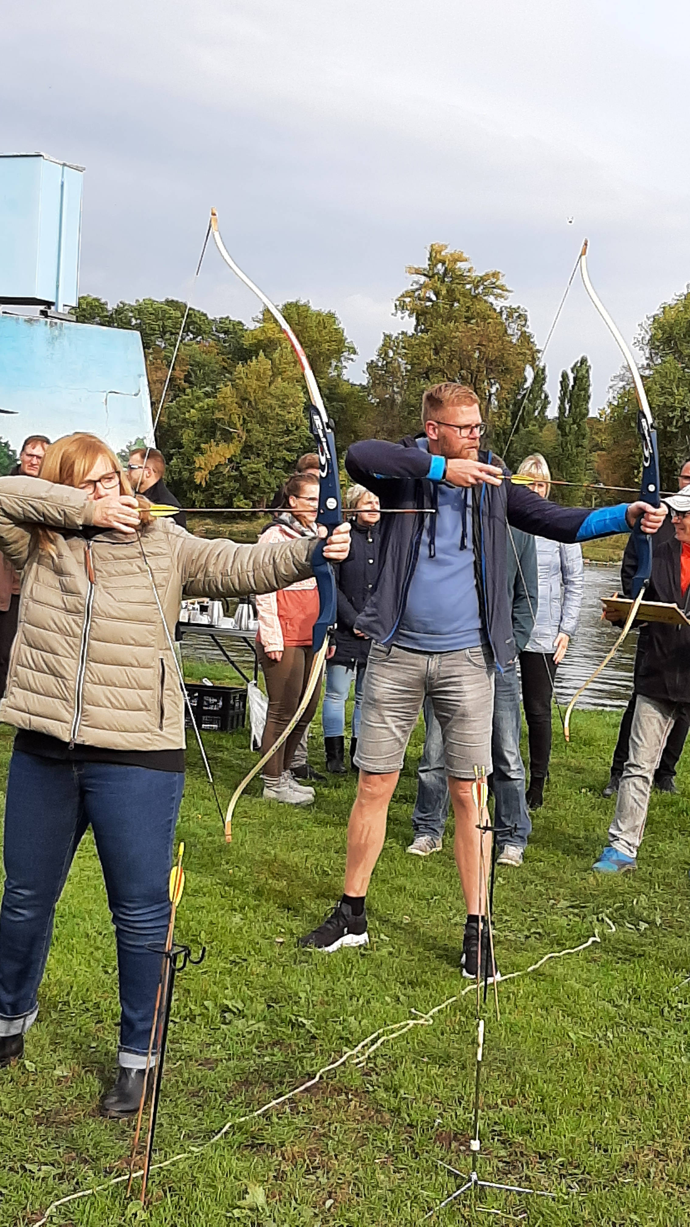 Erwachsene üben Bogenschießen auf einer Wiese, während umstehende Personen zusehen.