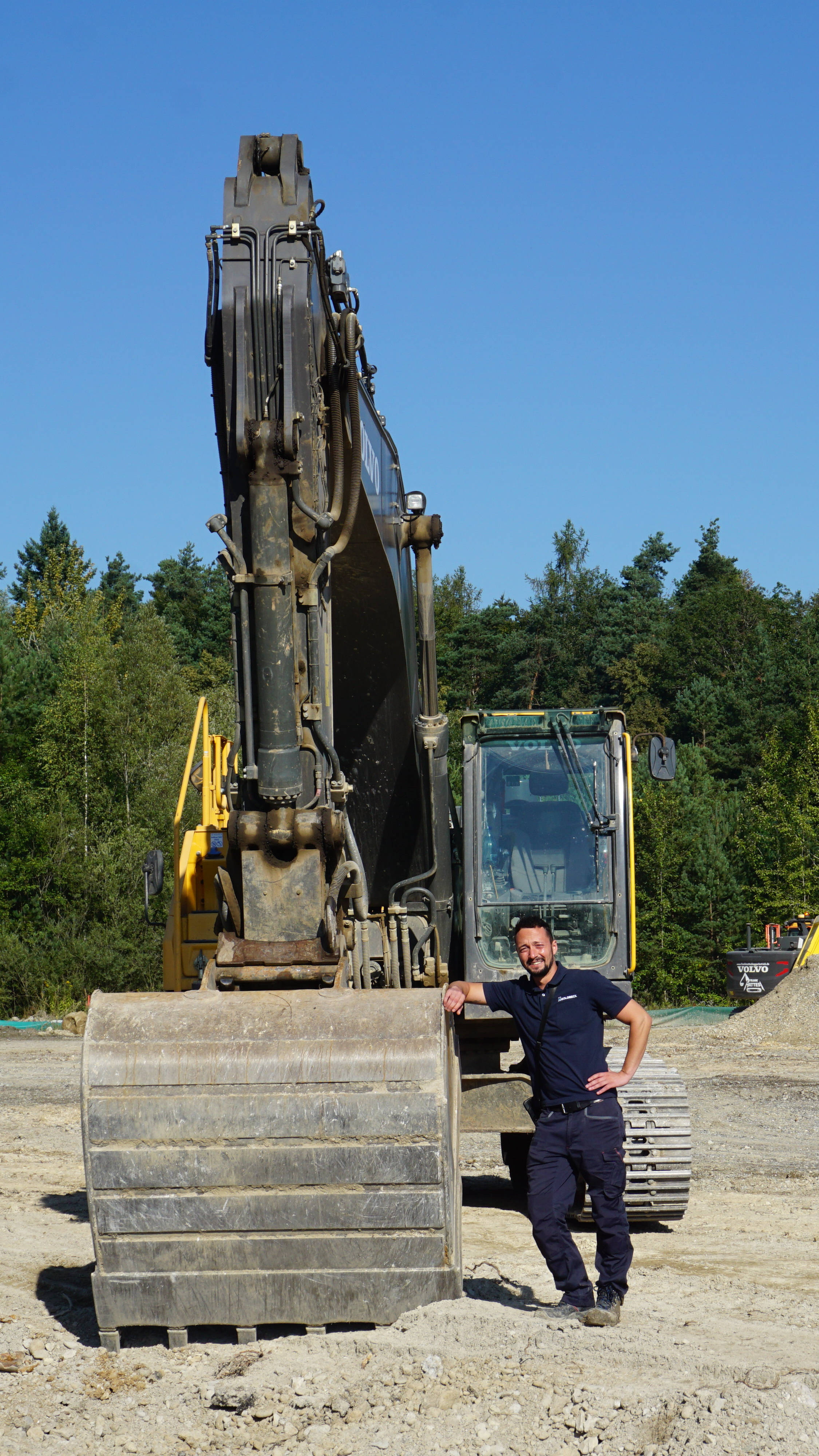 Bauarbeiter lehnt an einen großen Bagger auf einer Baustelle im Freien, umgeben von Wald und Kieshaufen.