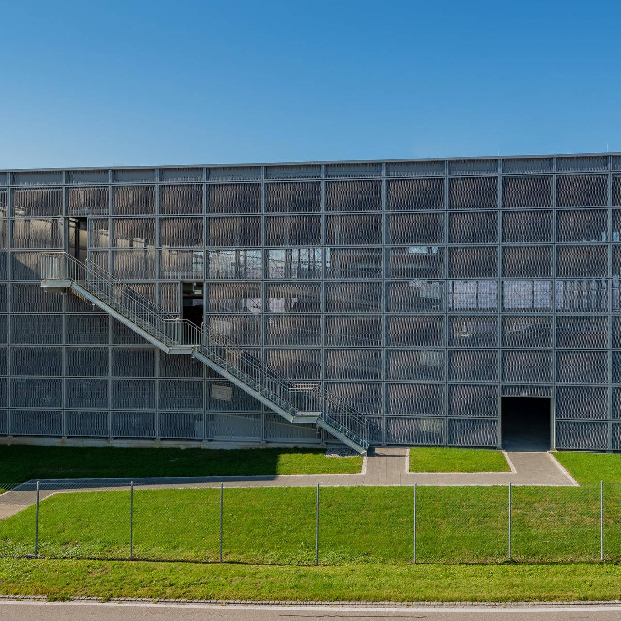 Mehrstöckige Parkgarage mit Treppenzugang und Metallfassade, umgeben von grünem Rasen und einem Zaun.