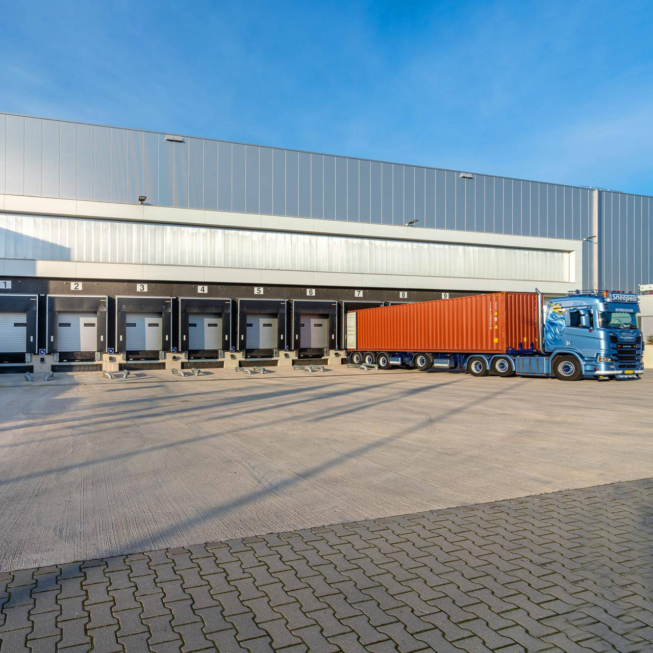 Vrachtwagen met blauwe cabine en oranje container geparkeerd bij laadperrons van een groot magazijn met de naam "flex" op de gevel.