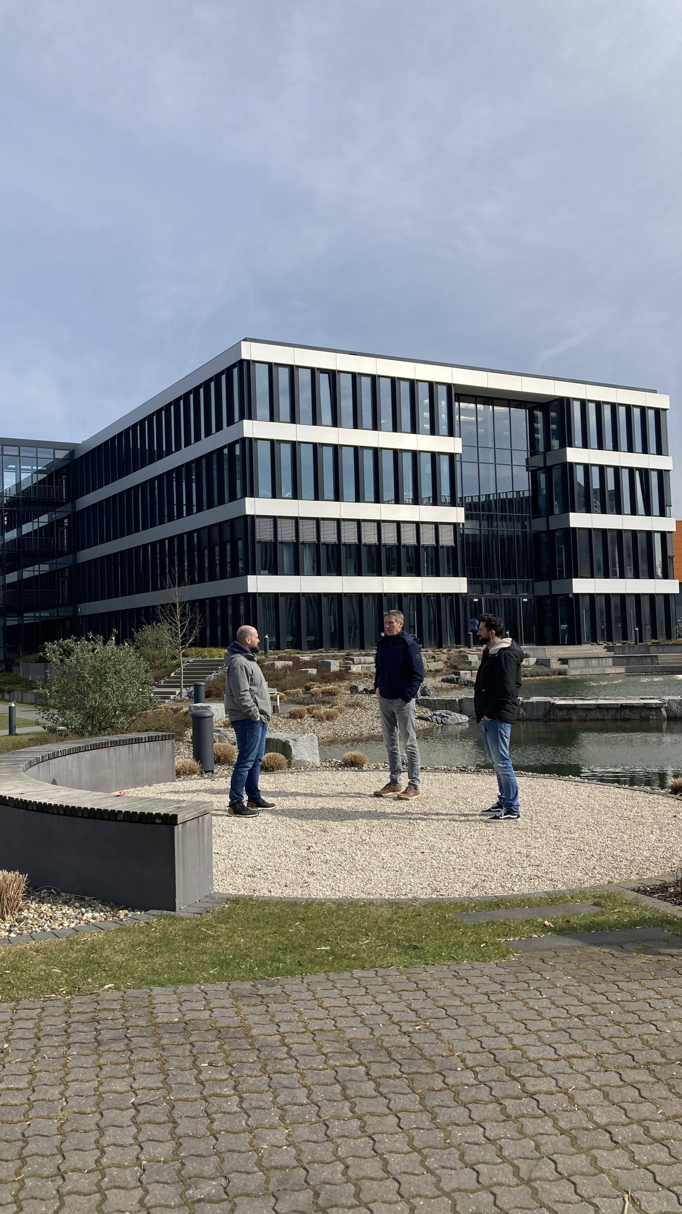 Modernes Bürogebäude mit Glasfassaden, drei Personen stehen im Dialog vor dem Gebäude, umgeben von Landschaftsgestaltung und Kiesflächen.