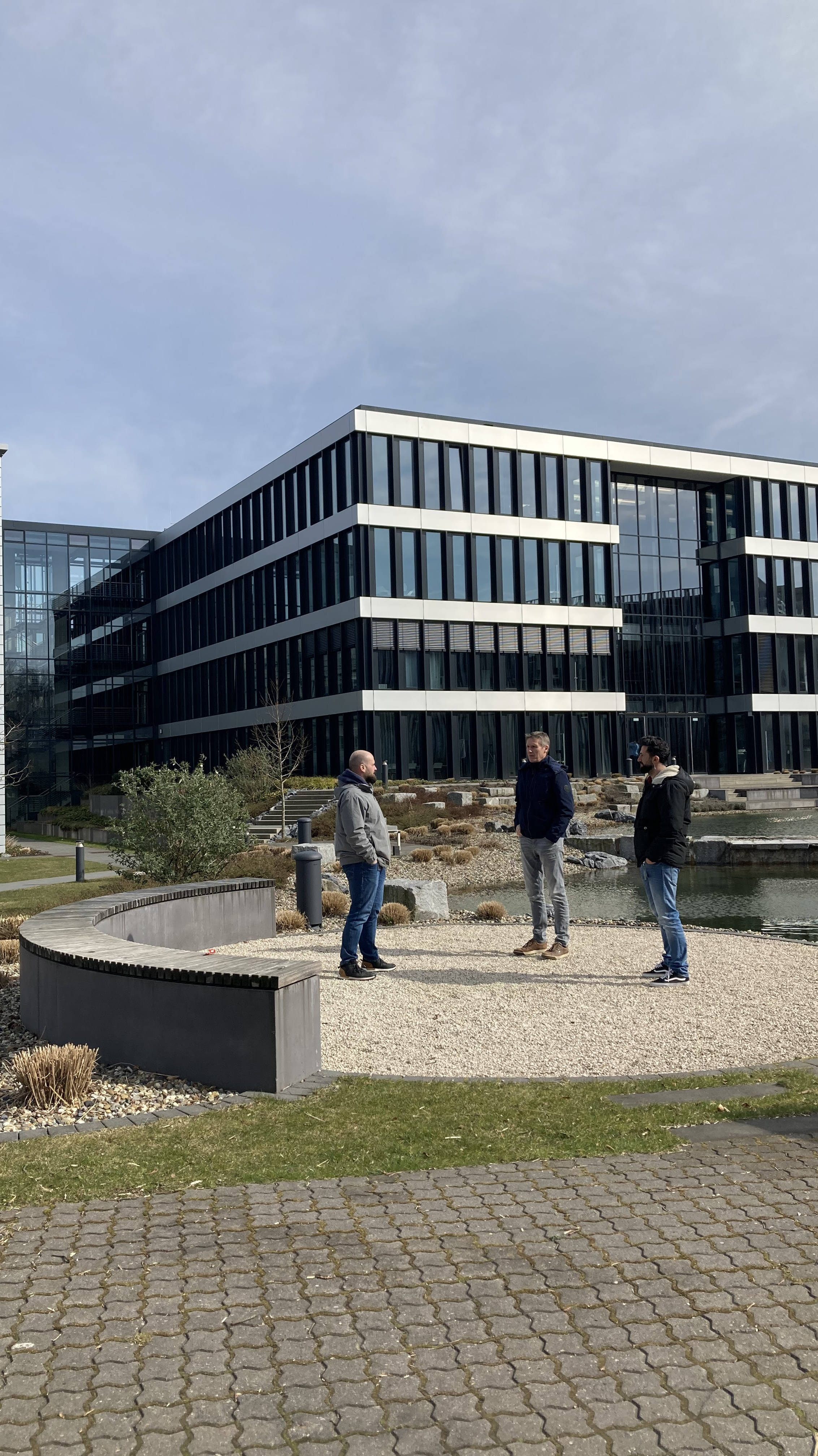 Modernes Bürogebäude mit Glasfassaden, drei Personen stehen im Dialog vor dem Gebäude, umgeben von Landschaftsgestaltung und Kiesflächen.