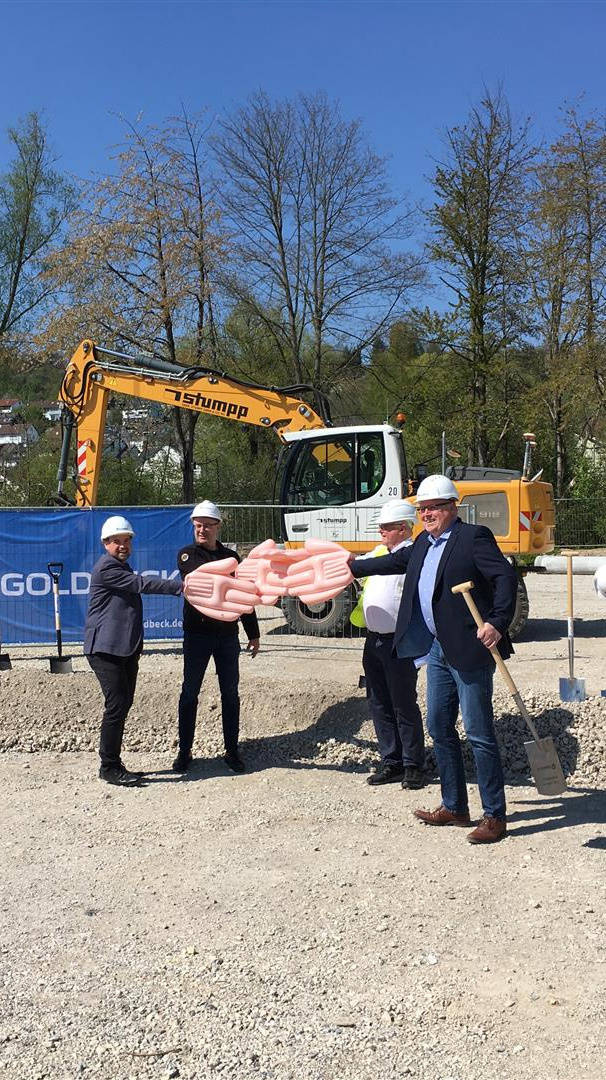 Personengruppe beim Spatenstich auf einer Baustelle, mit Baggern im Hintergrund und Bauplänen in der Hand.