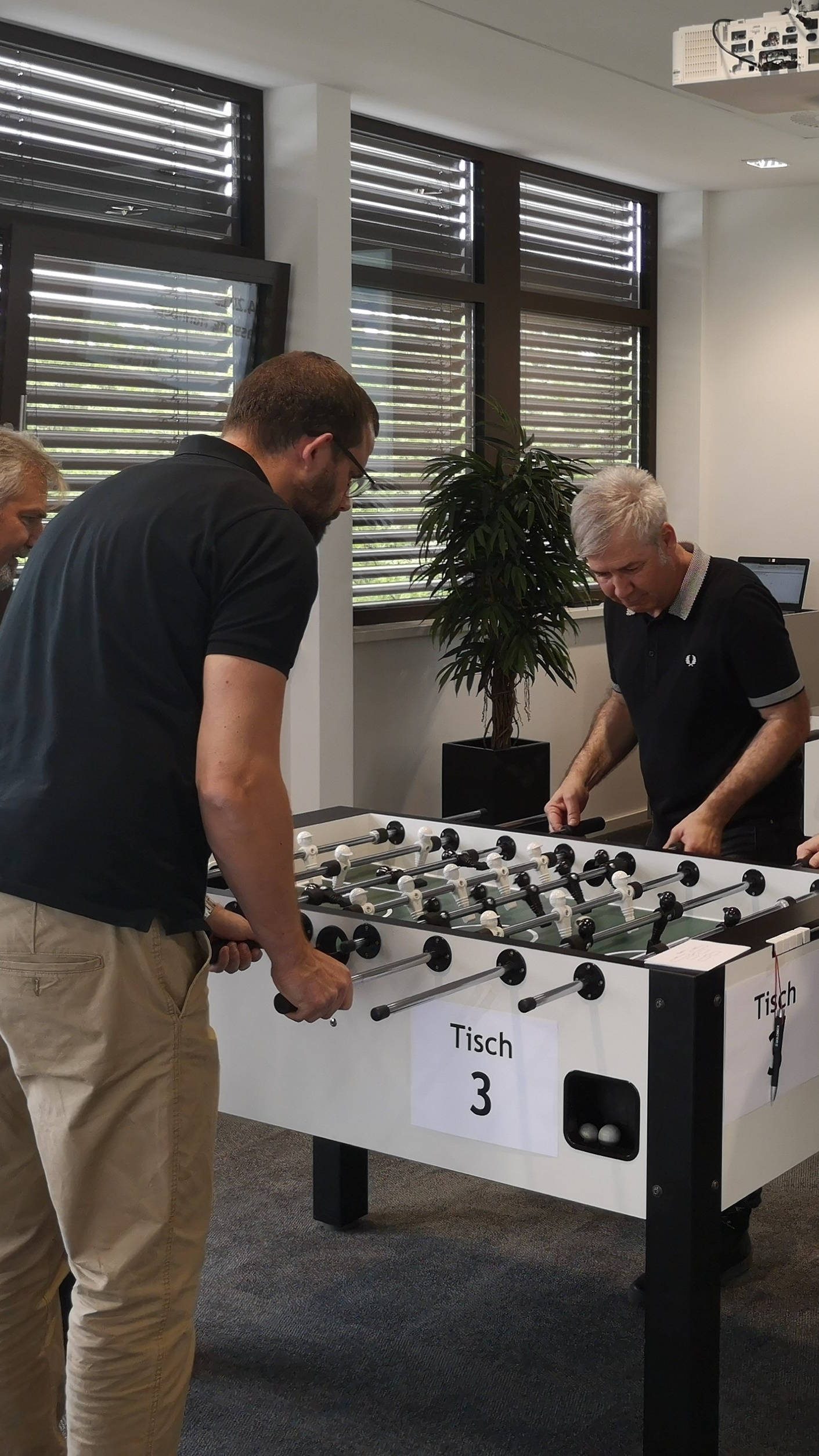 Four men playing table soccer in an office with large windows in the background.