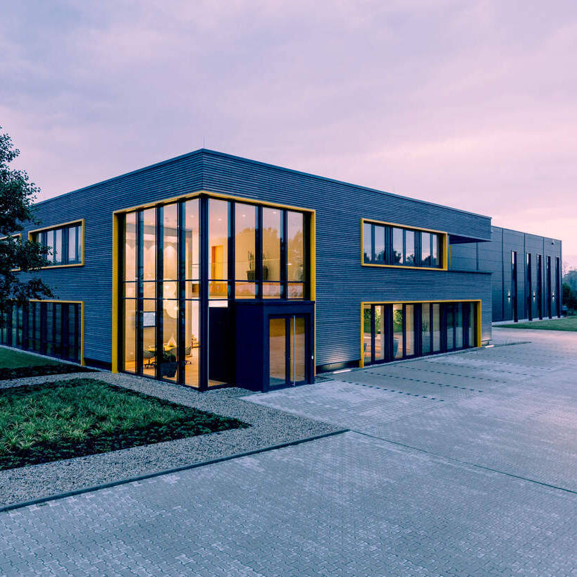 Modernes Bürogebäude mit großen Glasflächen und Parkplätzen im Vordergrund bei Abenddämmerung.