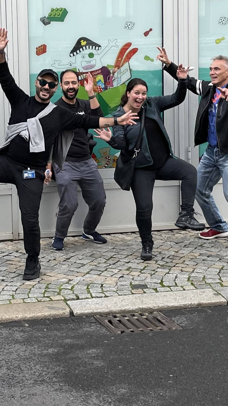 Four cheerful people pose in front of a colorful, child-friendly shop window decorated with drawings.