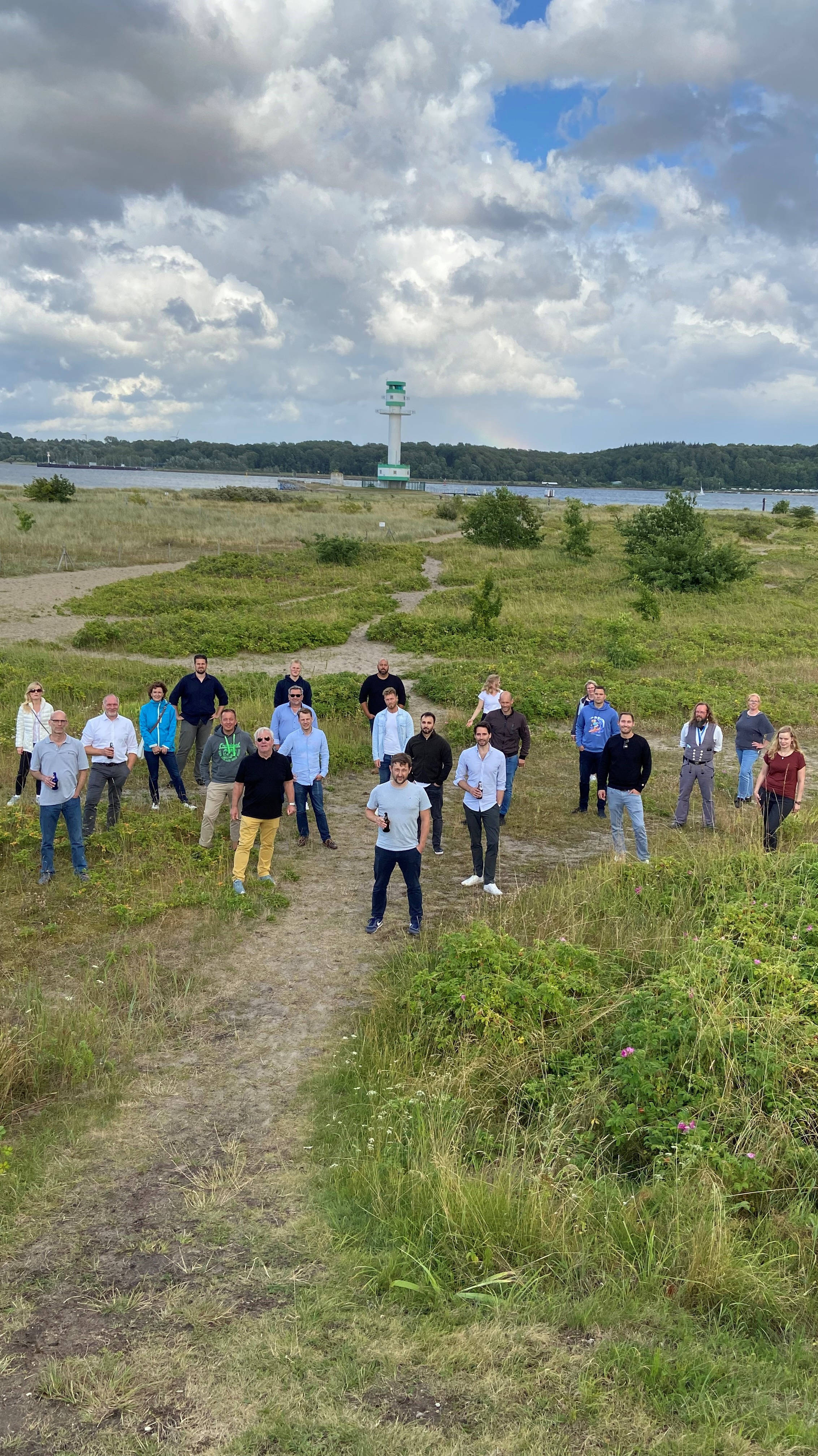 Gruppe von Menschen steht in einer grünen Küstenlandschaft mit einem Leuchtturm im Hintergrund unter bewölktem Himmel.