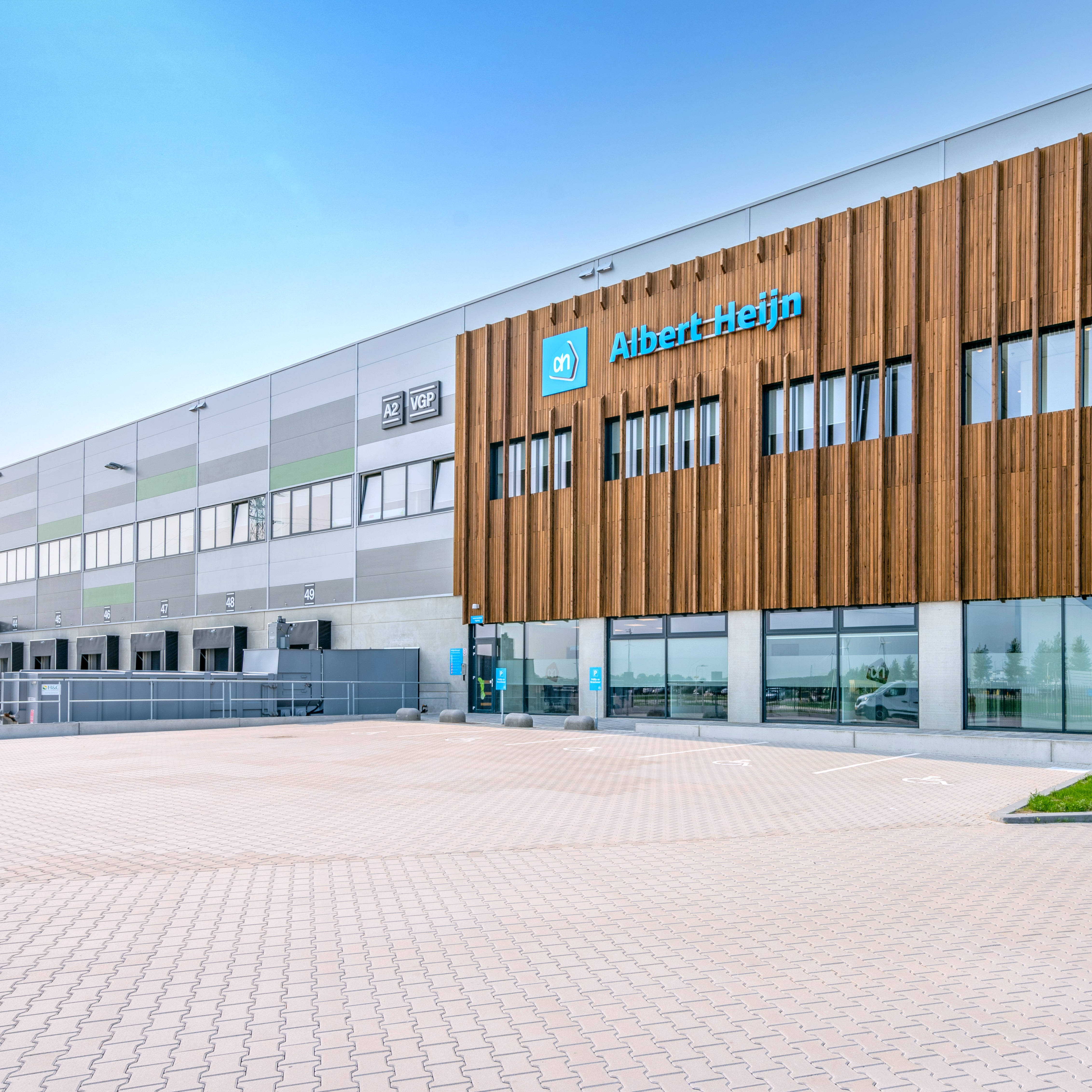 Moderne Lagerhalle mit einer Fassade, die sowohl aus Holzverkleidung als auch aus Glas- und Metallteilen besteht. Auf dem Gebäude ist der Name eines bekannten Einzelhandelsunternehmens zu sehen. Vor dem Eingang erstreckt sich ein großer, gepflasterter Parkplatz.