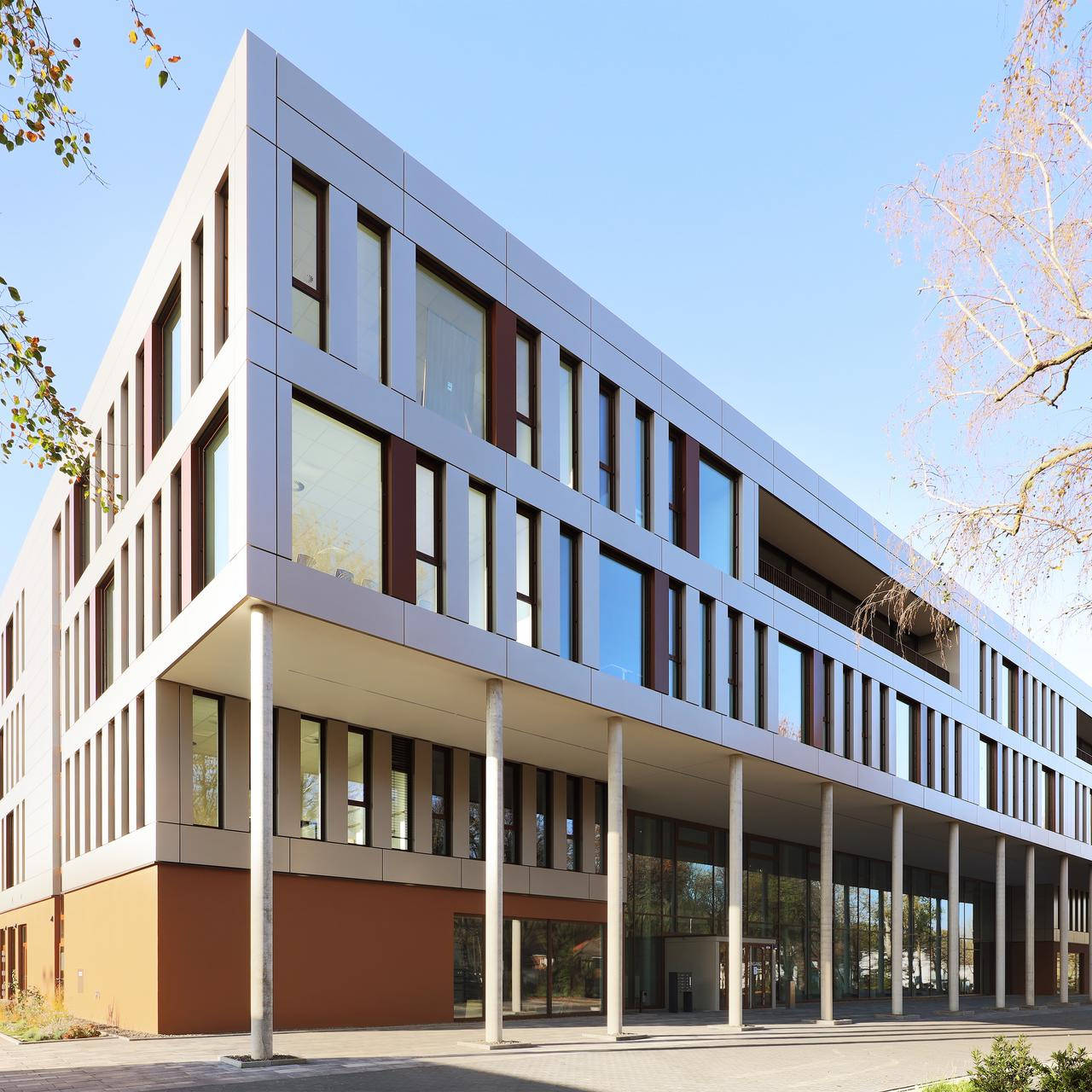 Modernes Bürogebäude mit großen Fenstern und klarer Architektur, umgeben von Bäumen an einem sonnigen Tag.