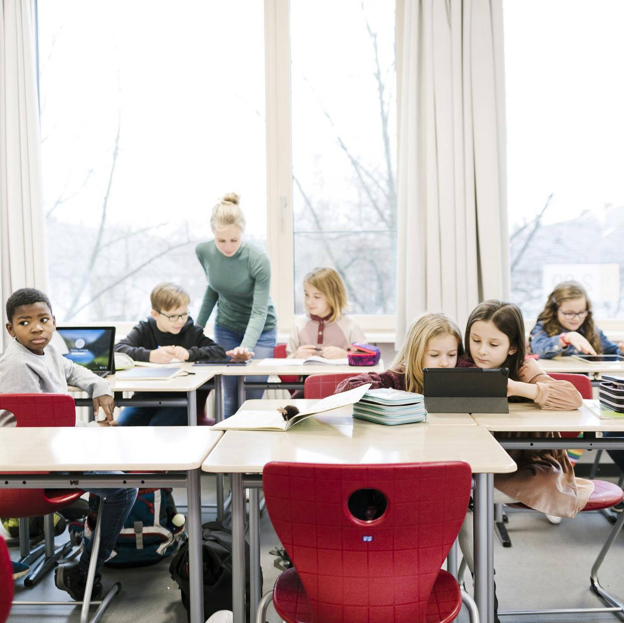 Primary school class in class, children sitting at tables, some using tablets, while a teacher leans over to them.