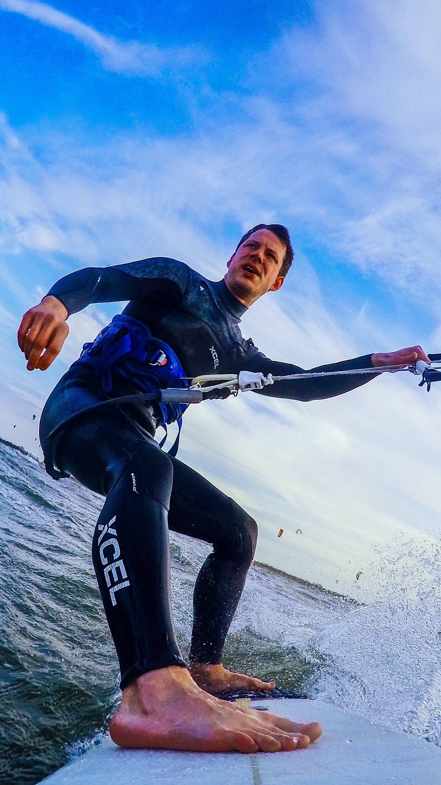 Kitesurfer in schwarz/blauem Neoprenanzug reitet über Wellen unter blauem Himmel, mit Blick auf eine weite Wasserfläche im Hintergrund.