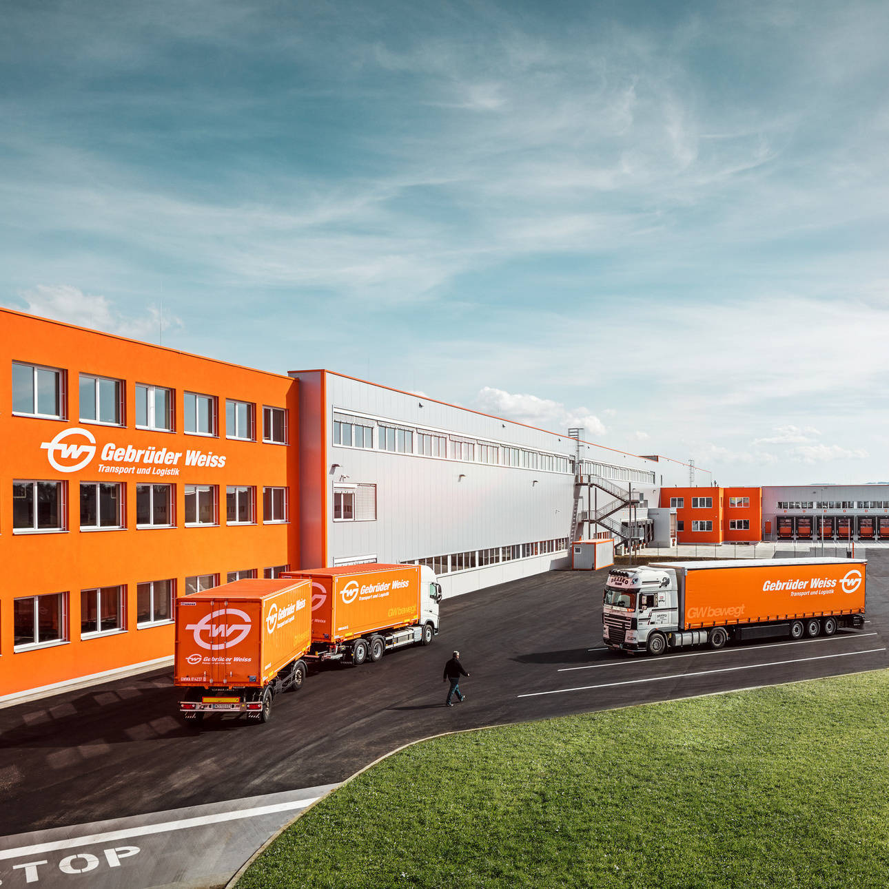Logistikzentrum mit mehreren orangefarbenen LKWs von Gebrüder Weiss vor einem Firmengebäude.