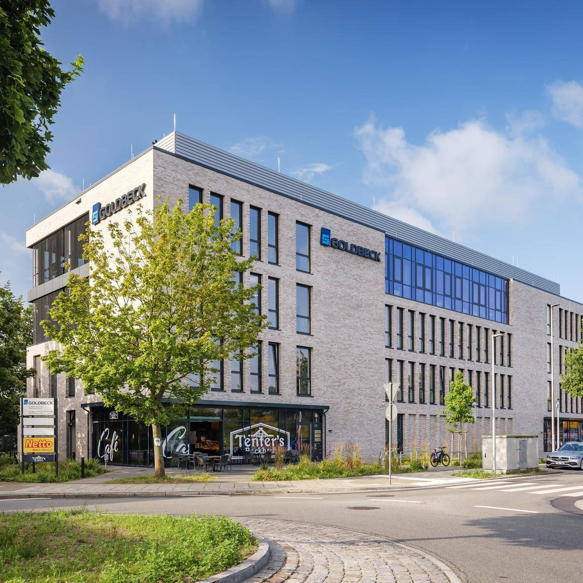 Modernes Bürogebäude mit dem Logo von Goldbeck, umgeben von Bäumen an einer ruhigen Straße bei klarem Himmel.