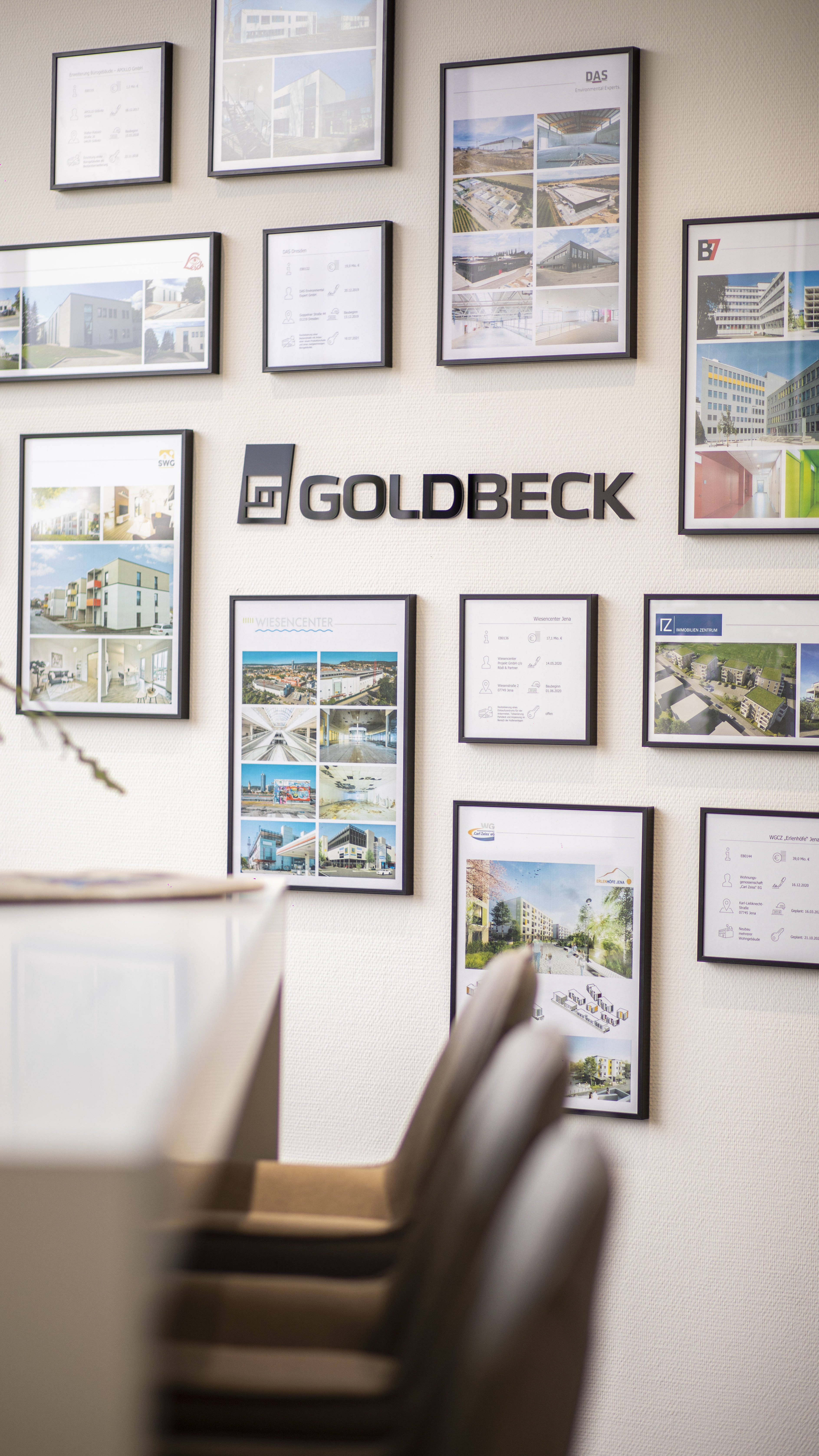 Wand mit gerahmten Architekturprojekten und Goldbeck-Logo, daneben Tisch und Stühle im Vordergrund.