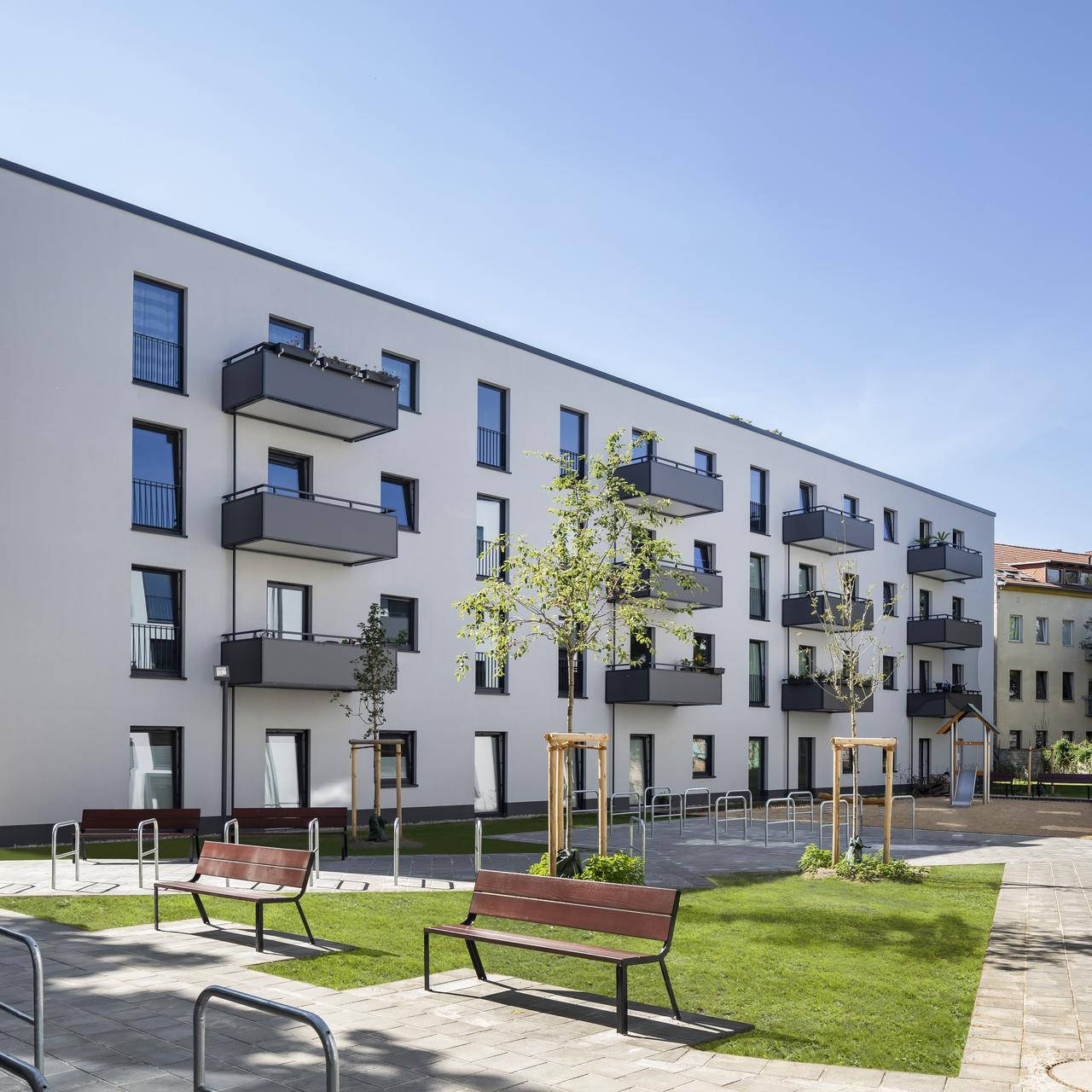 Modern residential complex with white facades and balconies, surrounded by a well-kept garden with benches and a small playground.