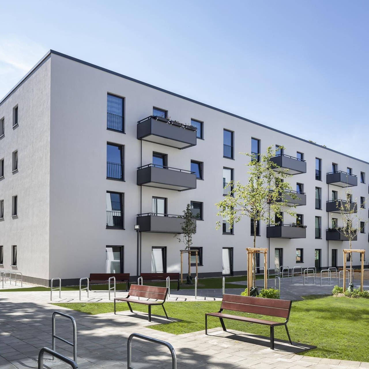 Modern residential complex with four-storey buildings, balconies, green inner courtyard and benches for sitting in sunny weather.
