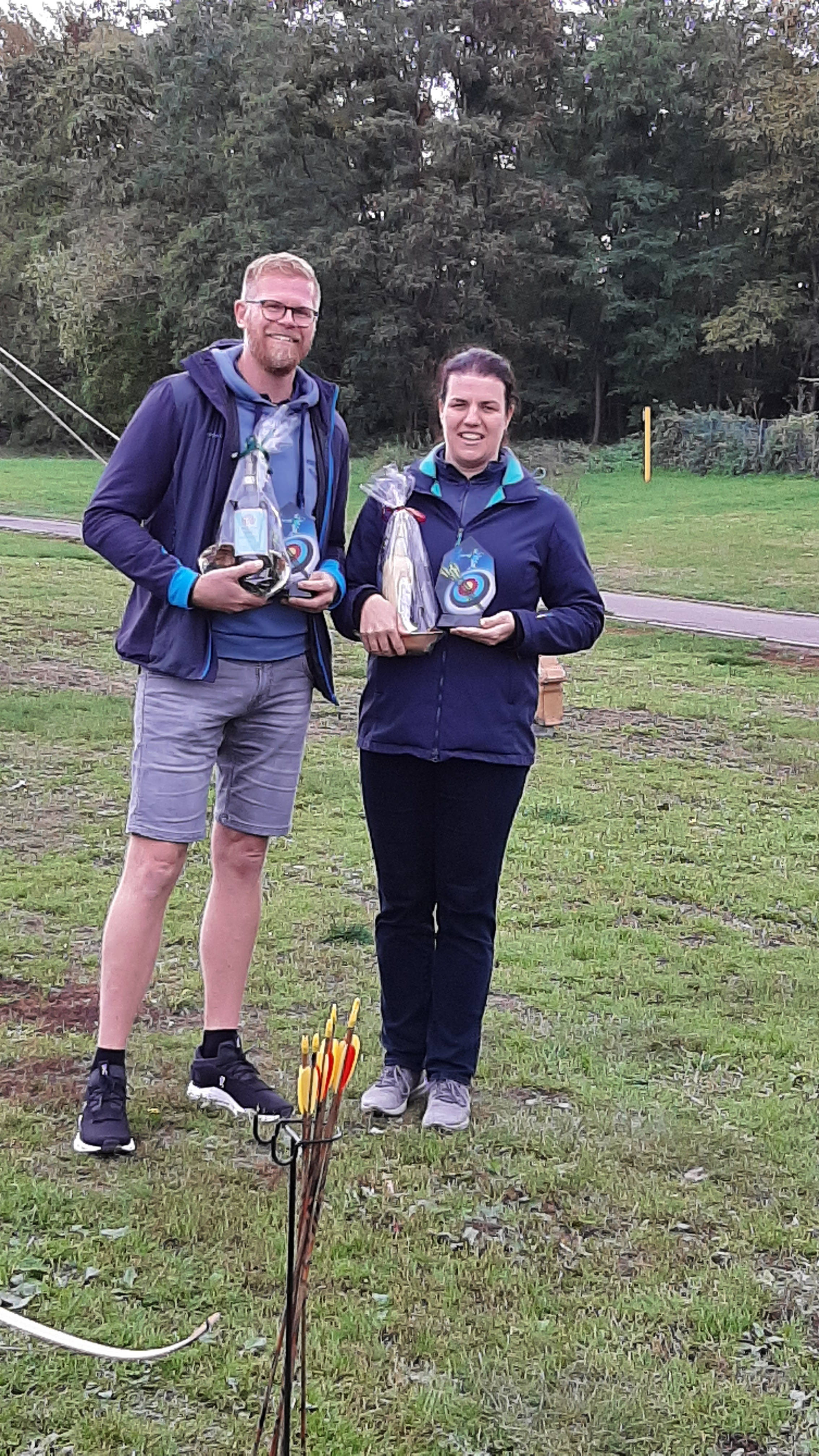 Zwei Personen stehen mit Glasflaschen und Geschenken auf einer Wiese vor einem bewaldeten Hintergrund, neben ihnen Bogensportausrüstung.