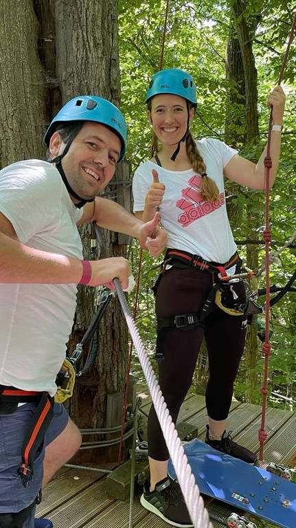 Zwei Personen mit Helmen und Sicherheitsgurten stehen lächelnd auf einer Plattform im Kletterpark, umgeben von Bäumen.