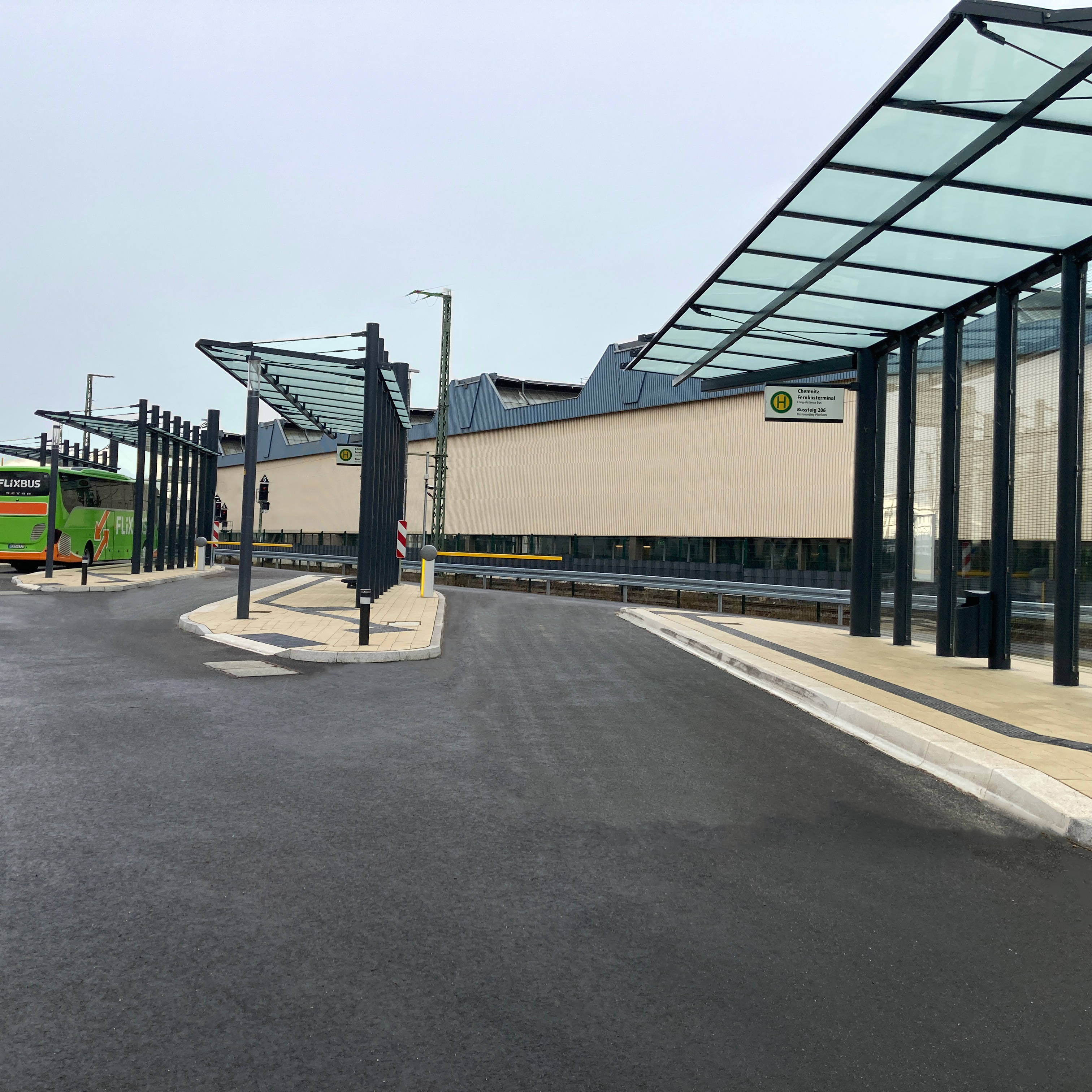 Moderner Busbahnhof mit grünem Reisebus unter überdachten Wartebereichen auf asphaltierter Fläche.