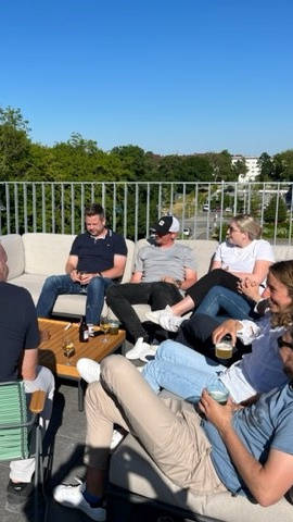 Eine Gruppe von Menschen sitzt bei sonnigem Wetter auf der Dachterrasse zusammen und unterhält sich.