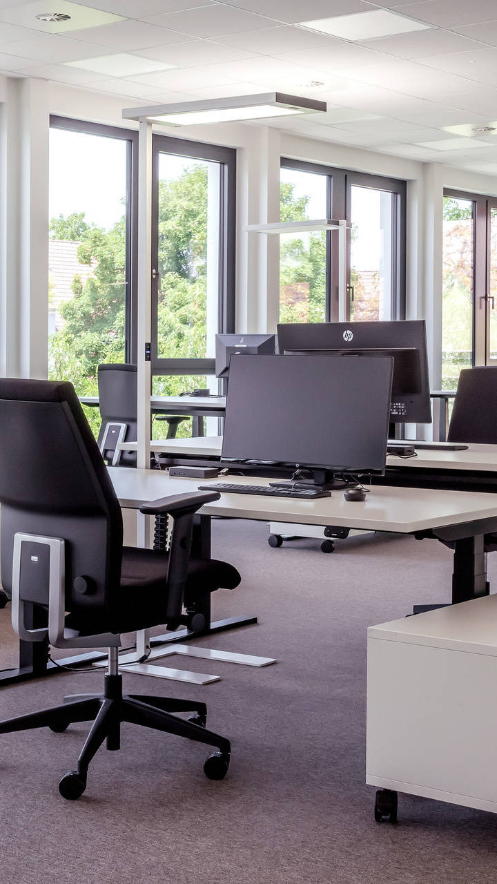 Moderner, heller Büroraum mit mehreren höhenverstellbaren Schreibtischen und ergonomischen Bürostühlen, umgeben von großen Fenstern.