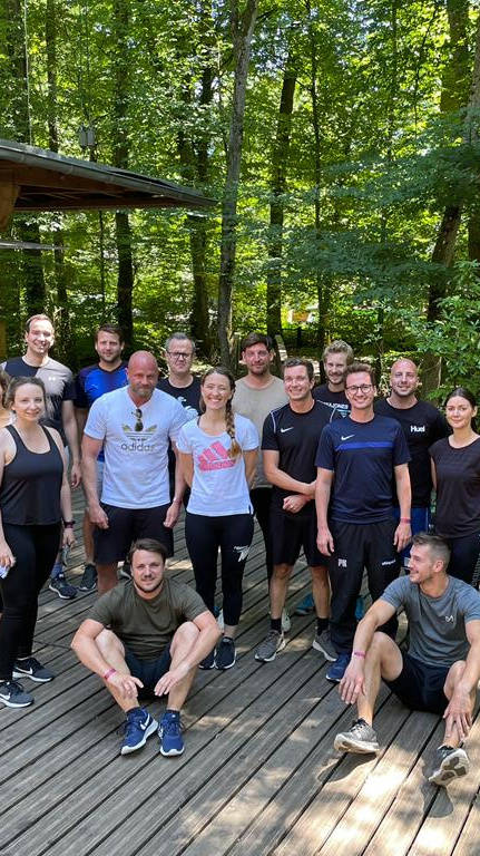 Gruppe von Menschen in Freizeitkleidung posiert auf einer hölzernen Plattform im Wald, nahe einem Kassenhaus und einem Kletterparcours.