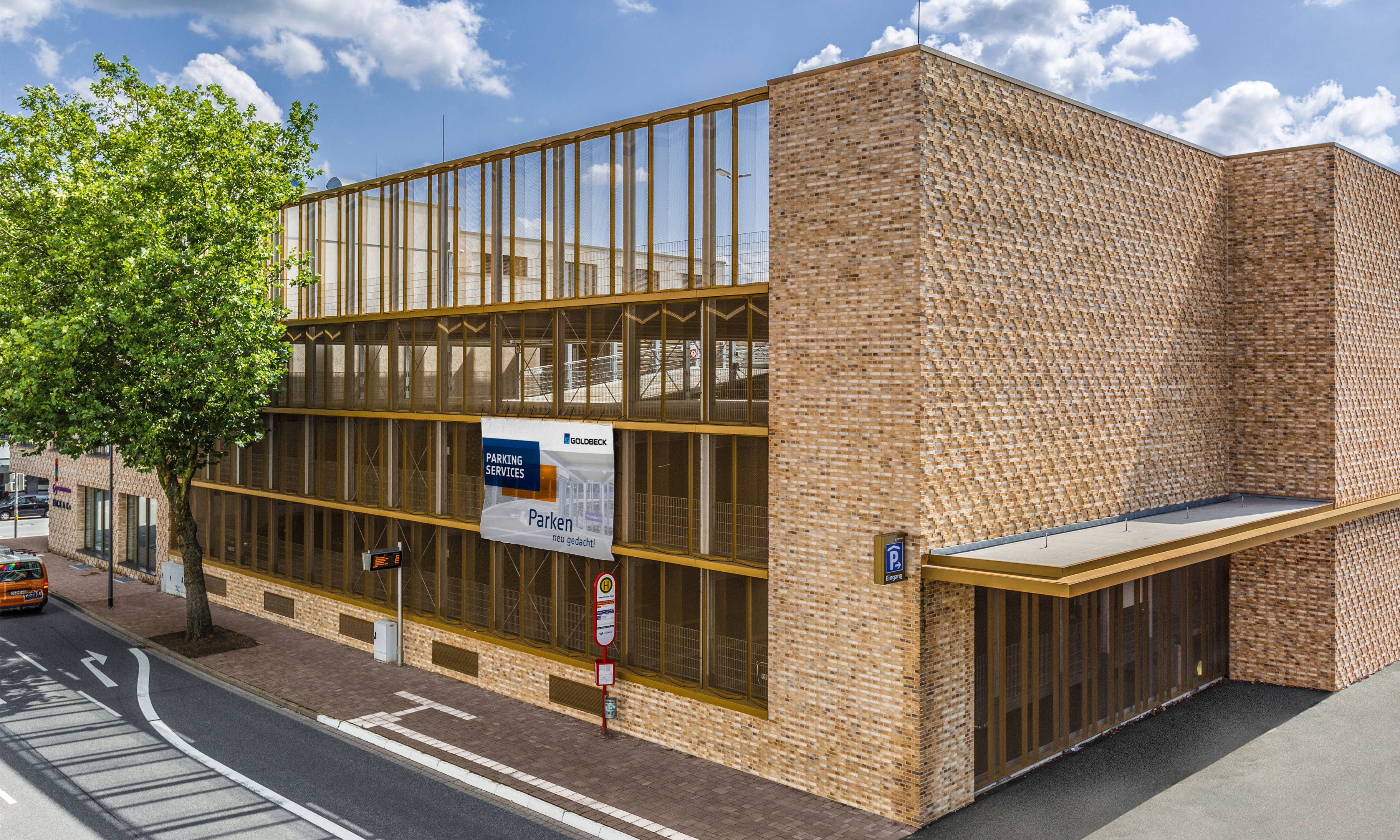 Modern parking garage with brick facade and glass facade, next to it a tree and a signpost for parking services.