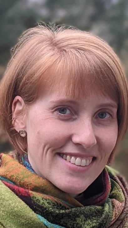 Eine lächelnde Frau mit kurzen roten Haaren trägt einen bunten Schal, im Hintergrund Natur.
