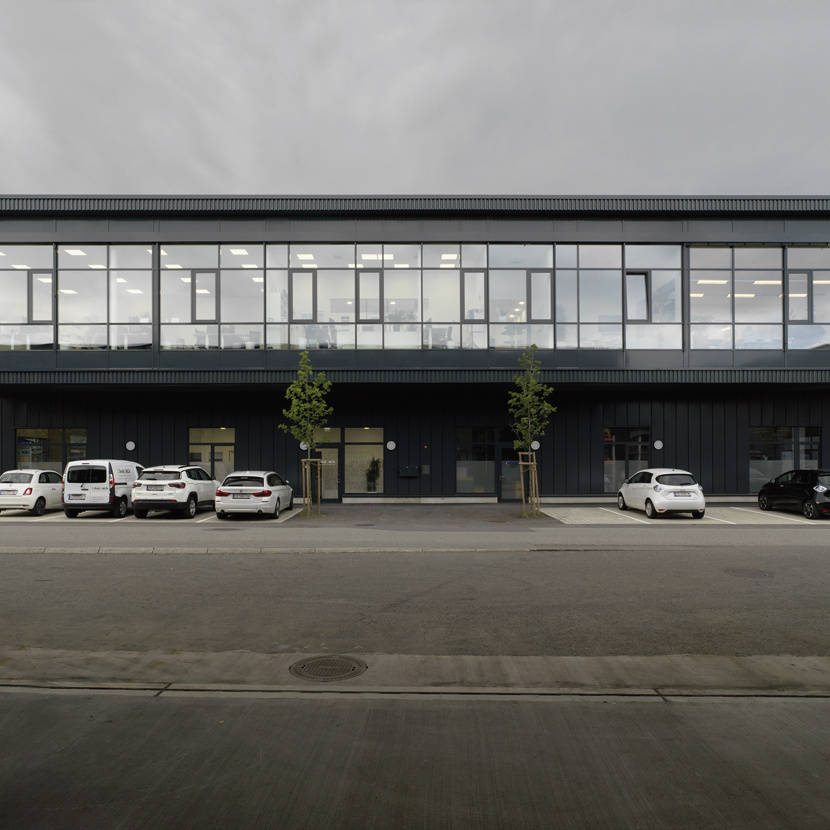 Modernes zweistöckiges Bürogebäude mit großen Fenstern, parkende Autos davor, bedeckter Himmel.