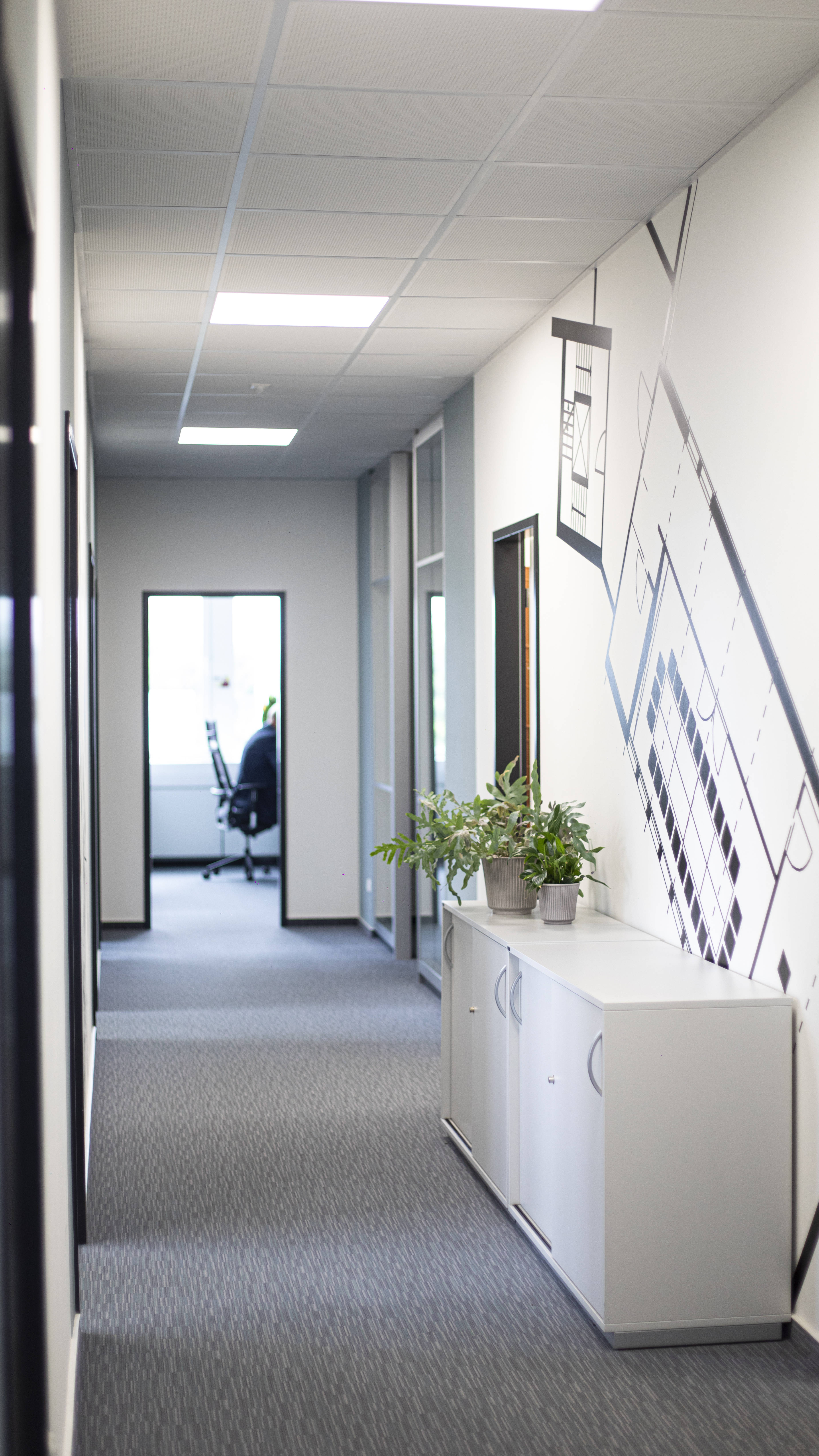 Moderner Büroflur mit Teppichboden, weißen Schränken, Pflanzen und Wandgrafik, im Hintergrund ein Schreibtisch mit Stuhl.