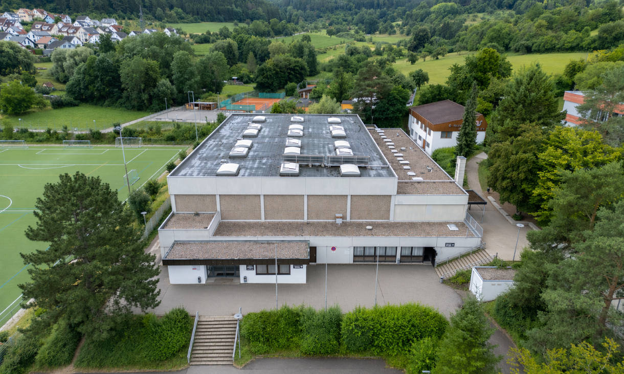 A modern gymnasium in a rural setting, with surrounding sports fields and residential buildings in the background.