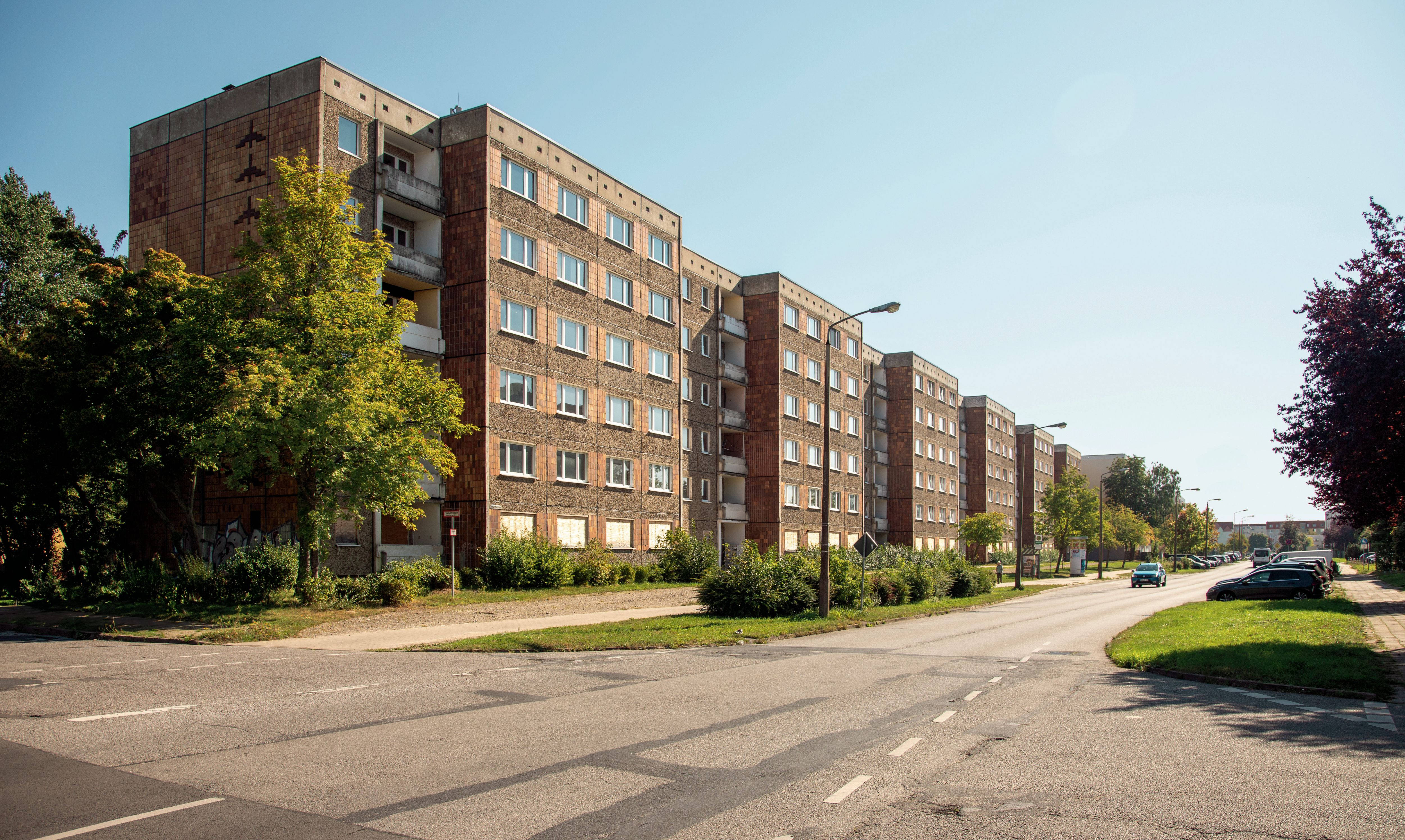Multi-storey brick block of flats in need of renovation on a sunny street with parked cars and trees.