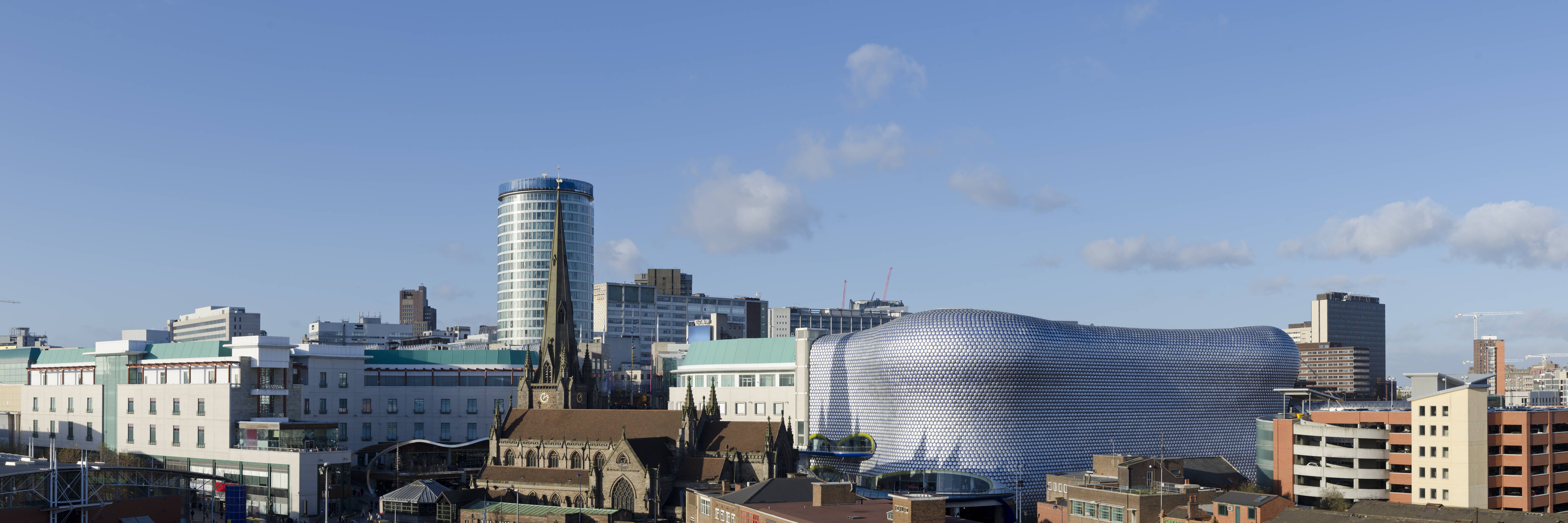 Stadtpanorama von Birmingham mit modernem Einkaufszentrum und historischer Kirche unter blauem Himmel.