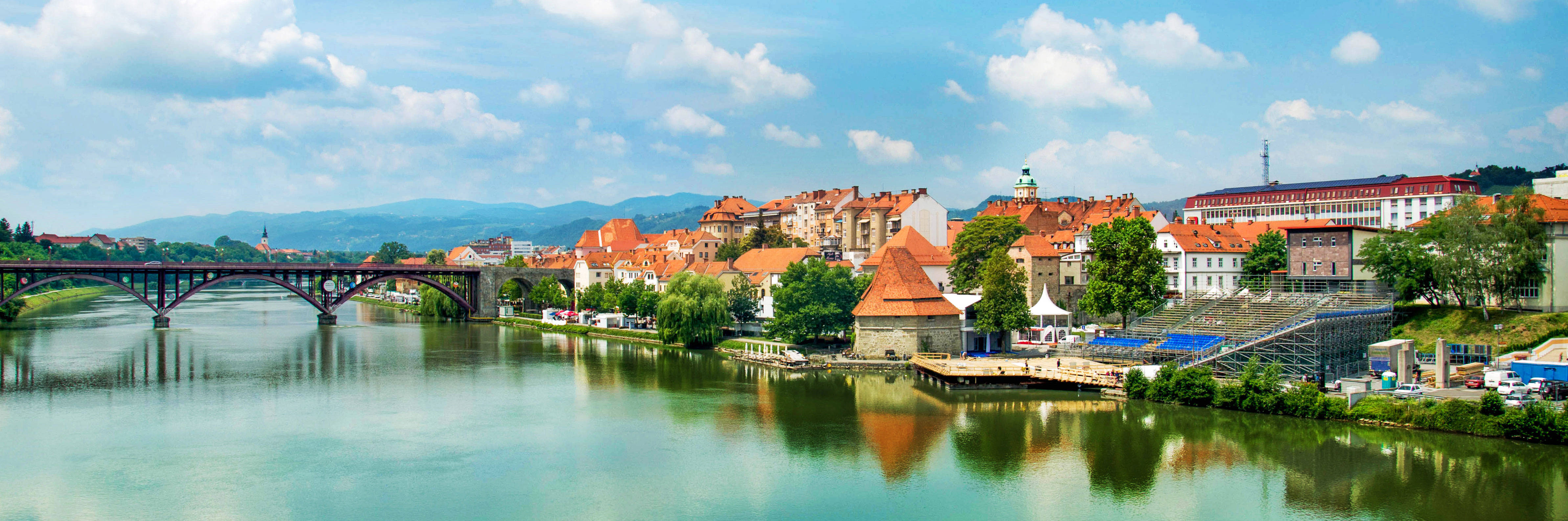 Maribor-Panorama mit alter Brücke über einen Fluss, historischen Gebäuden und grünen Hügeln im Hintergrund unter blauem Himmel.