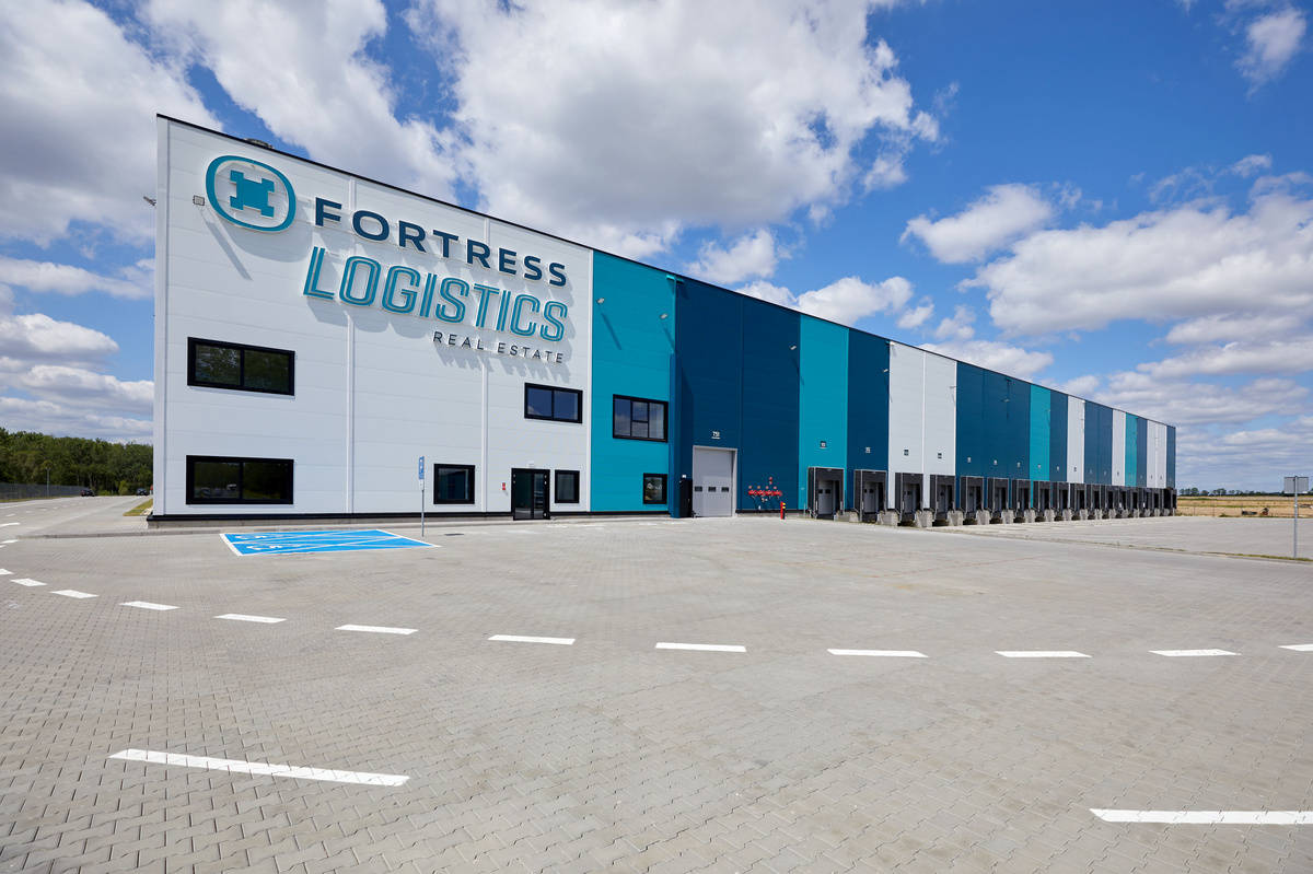 Duży magazyn logistyczny o nowoczesnym designie z oznaczeniami "FORTRESS LOGISTICS REAL ESTATE" na białej i niebieskiej fasadzie. Przed budynkiem znajduje się rozległy, brukowany plac, a w tle widać niebo z rozproszonymi chmurami.