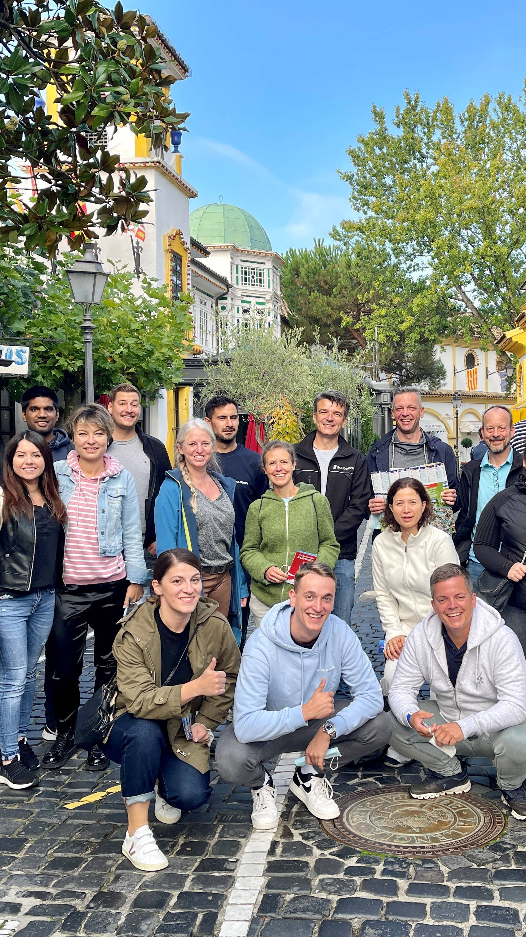 Gruppe von Menschen steht auf gepflasterter Straße vor bunt verzierten Gebäuden. Sie lächeln und posieren entspannt für ein Gruppenfoto.