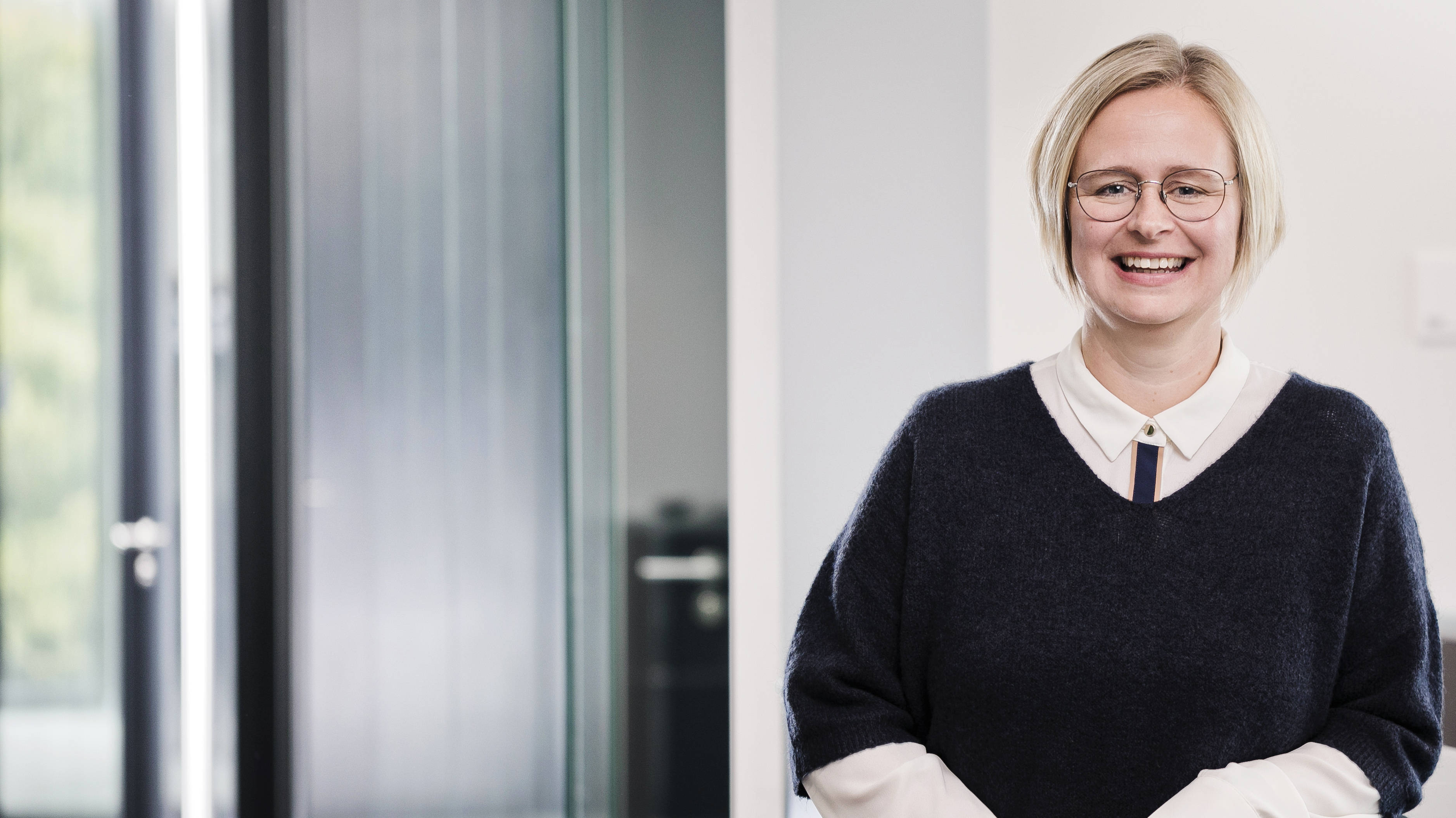 portraitfoto einer frau mit kurzen blonden haaren und einer brille steht angelehnt an einer fläche