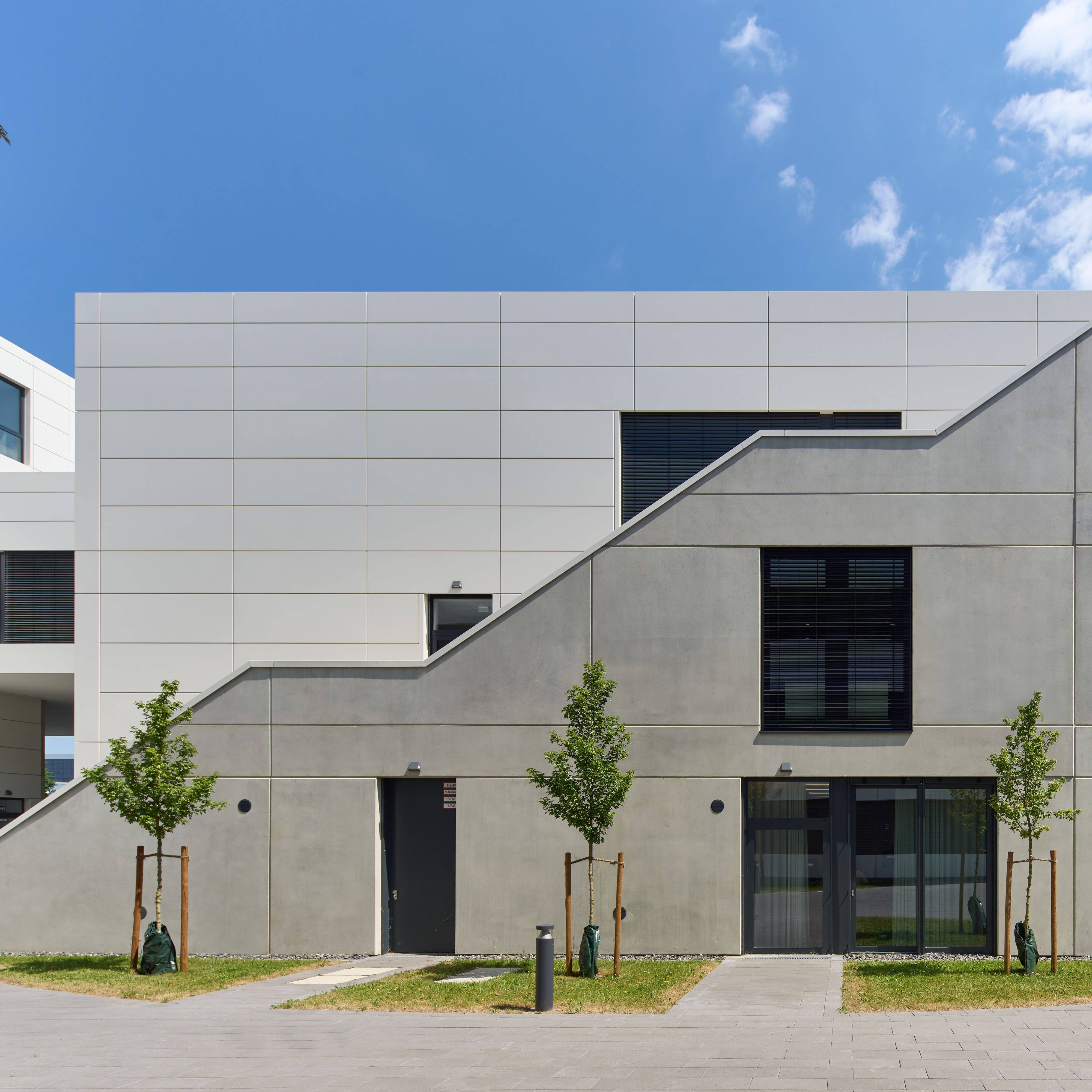 Modern architecture of a white building with geometric shapes, small windows and young trees in the foreground.