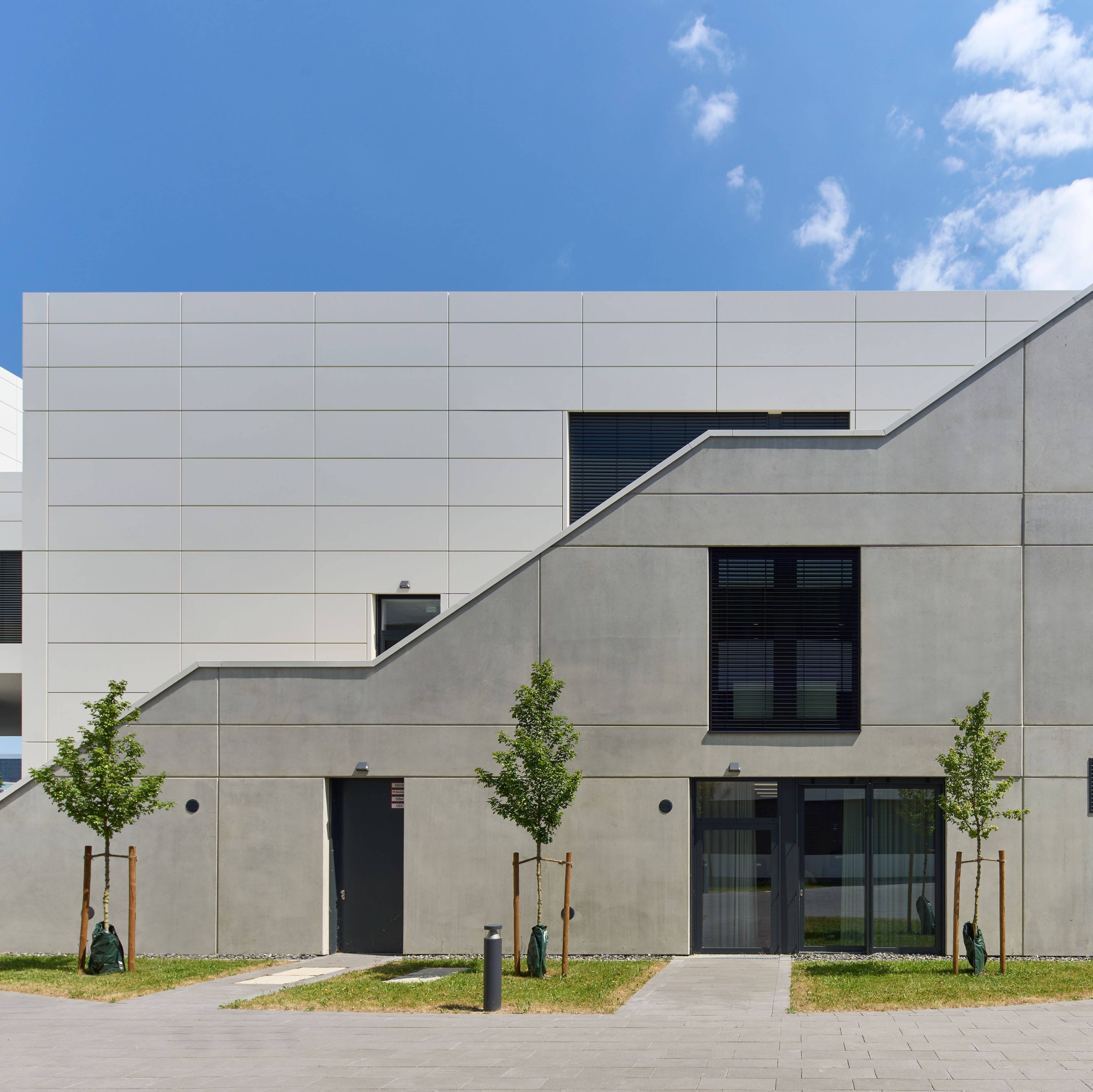 Moderne Architektur eines weißen Gebäudes mit geometrischen Formen, kleinen Fenstern und jungen Bäumen im Vordergrund.