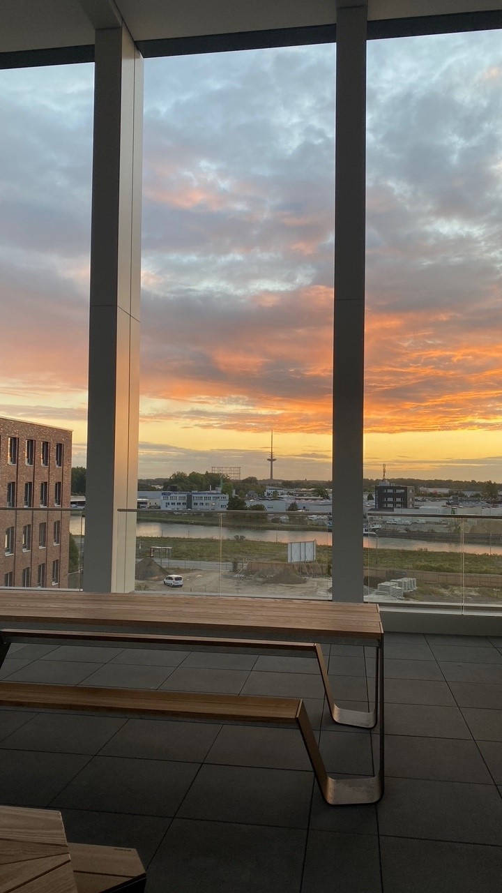 Blick durch große Fenster auf einen Sonnenuntergang über einer Stadtlandschaft, im Vordergrund Holztische.