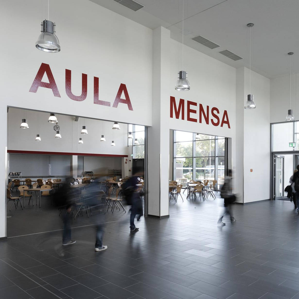 Modern assembly hall and canteen entrance area with people walking, hanging lamps and large windows.