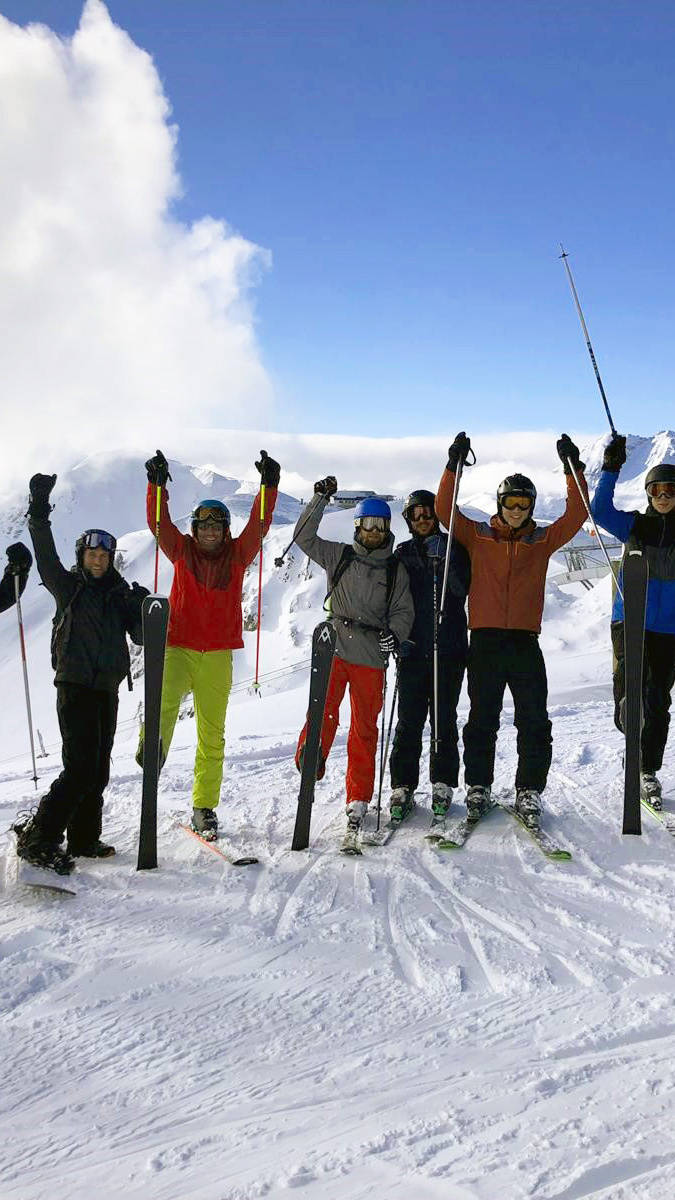 Gruppe von Skifahrern in bunter Kleidung hebt jubelnd die Arme auf einem schneebedeckten Berg vor blauem Himmel.