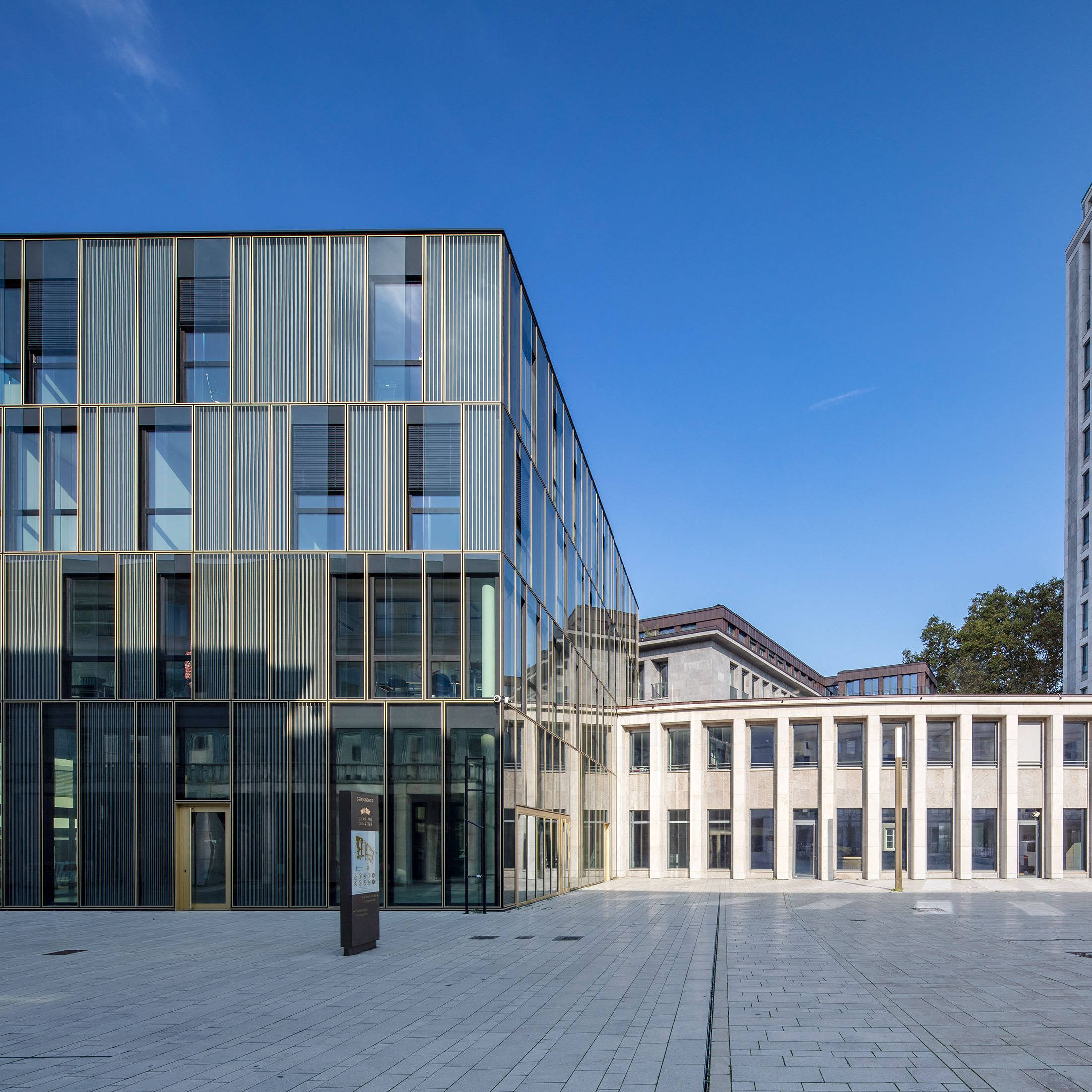 Modernes, gläsernes Bürogebäude mit vertikalen Metallstreifen, umgeben von einem gepflasterten Platz und klarem blauen Himmel.