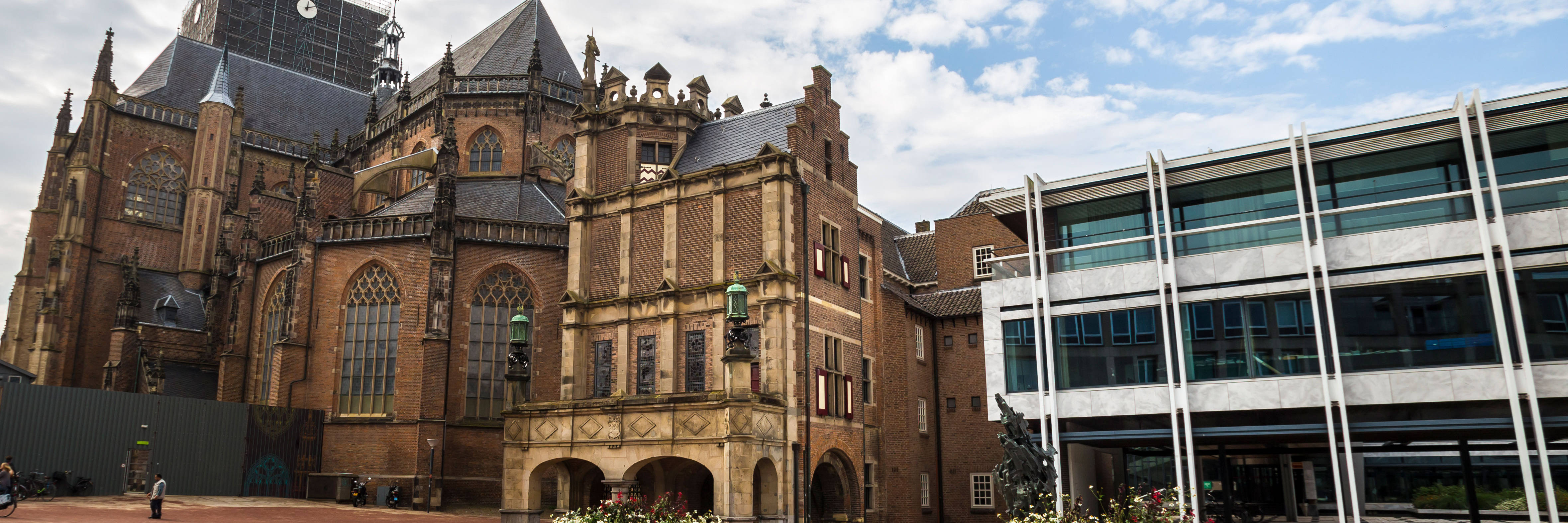 Large, old cathedral next to a modern building, with a paved forecourt and flower pots, under a cloudy sky.