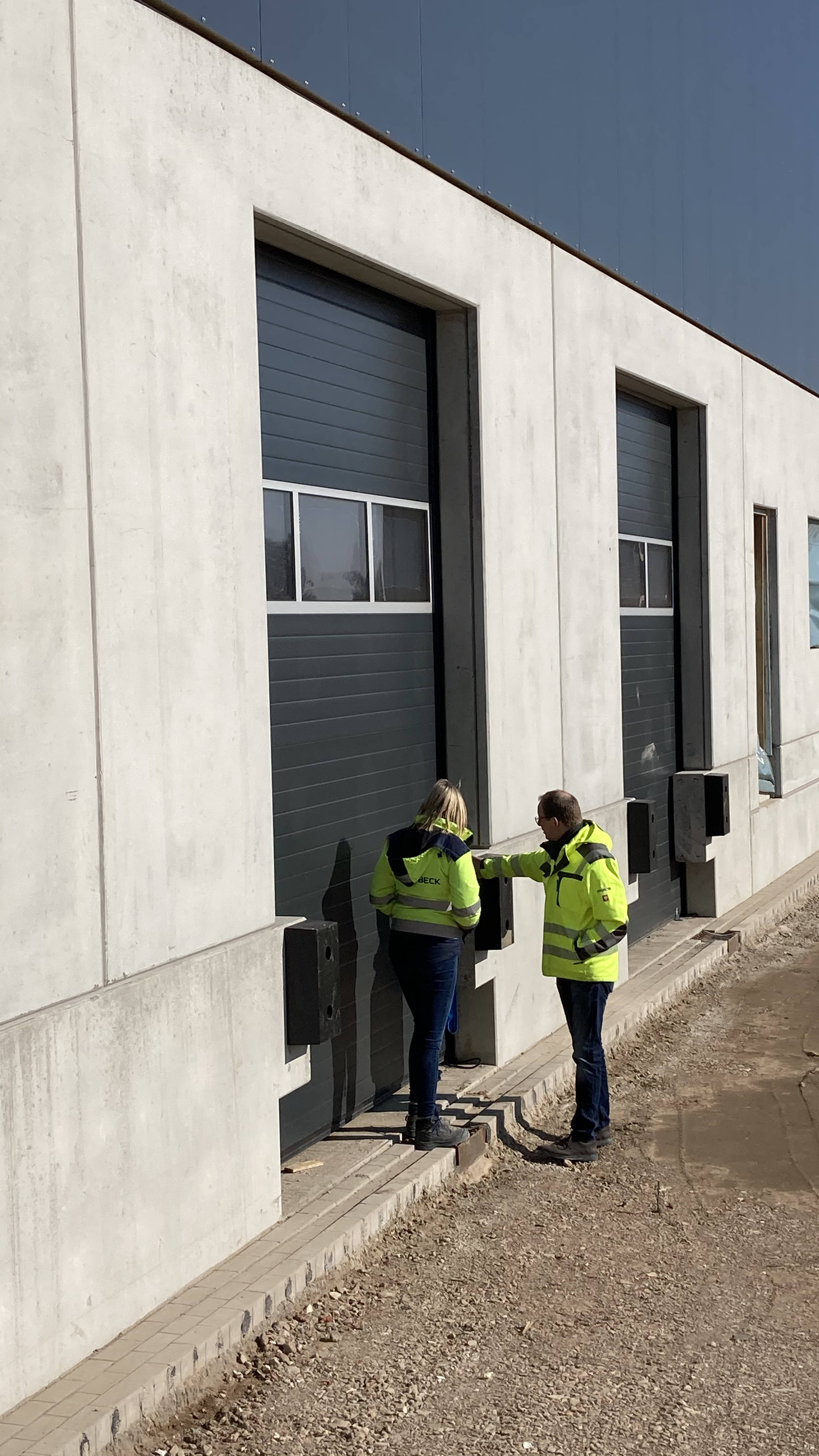 Zwei Personen in Warnwesten diskutieren vor einem Industriegebäudeeingang.
