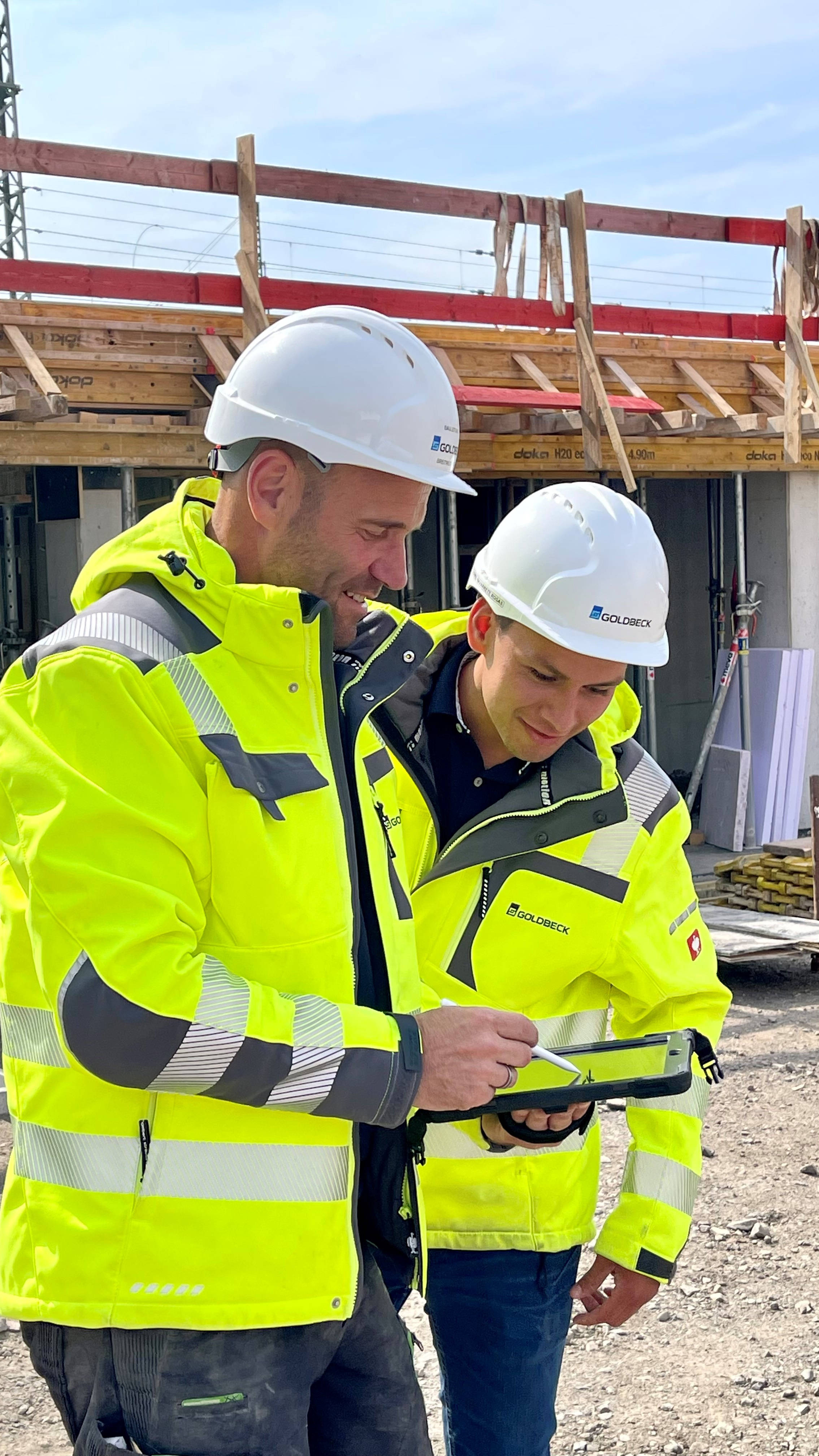Bauarbeiter in gelben Schutzwesten und Helmen betrachten gemeinsam ein Tablet auf einer Baustelle.