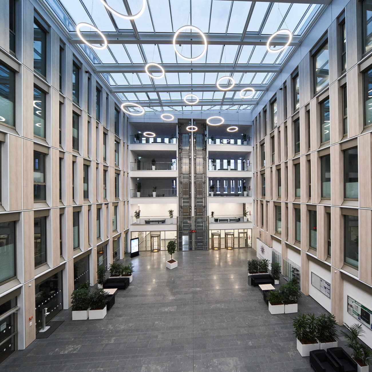 Modernes, lichtdurchflutetes Atrium eines Bürogebäudes mit Glasdach, hängenden Leuchtringen und begrünten Sitzbereichen.
